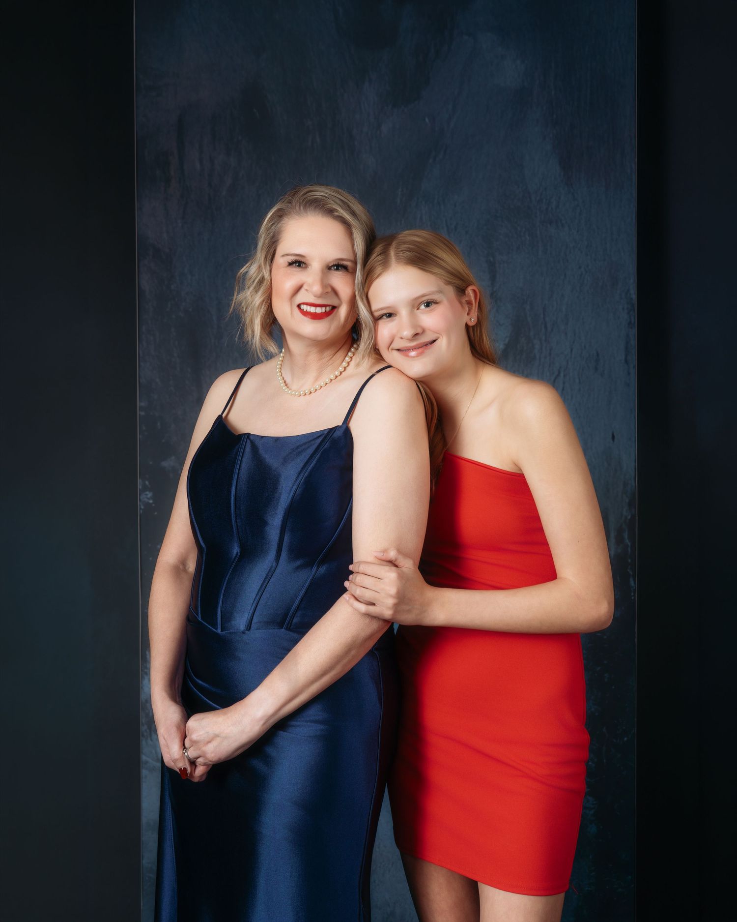 Two women in elegant red and navy dresses pose together smiling against a dark blue backdrop in a professional portrait.