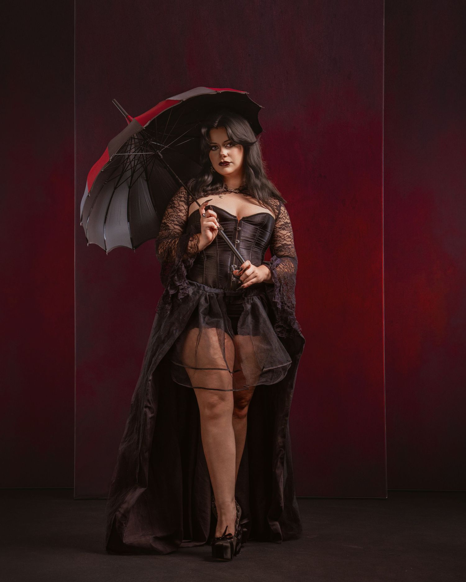 Gothic woman in black corset and lace coat holding a dark umbrella against a dramatic red background.