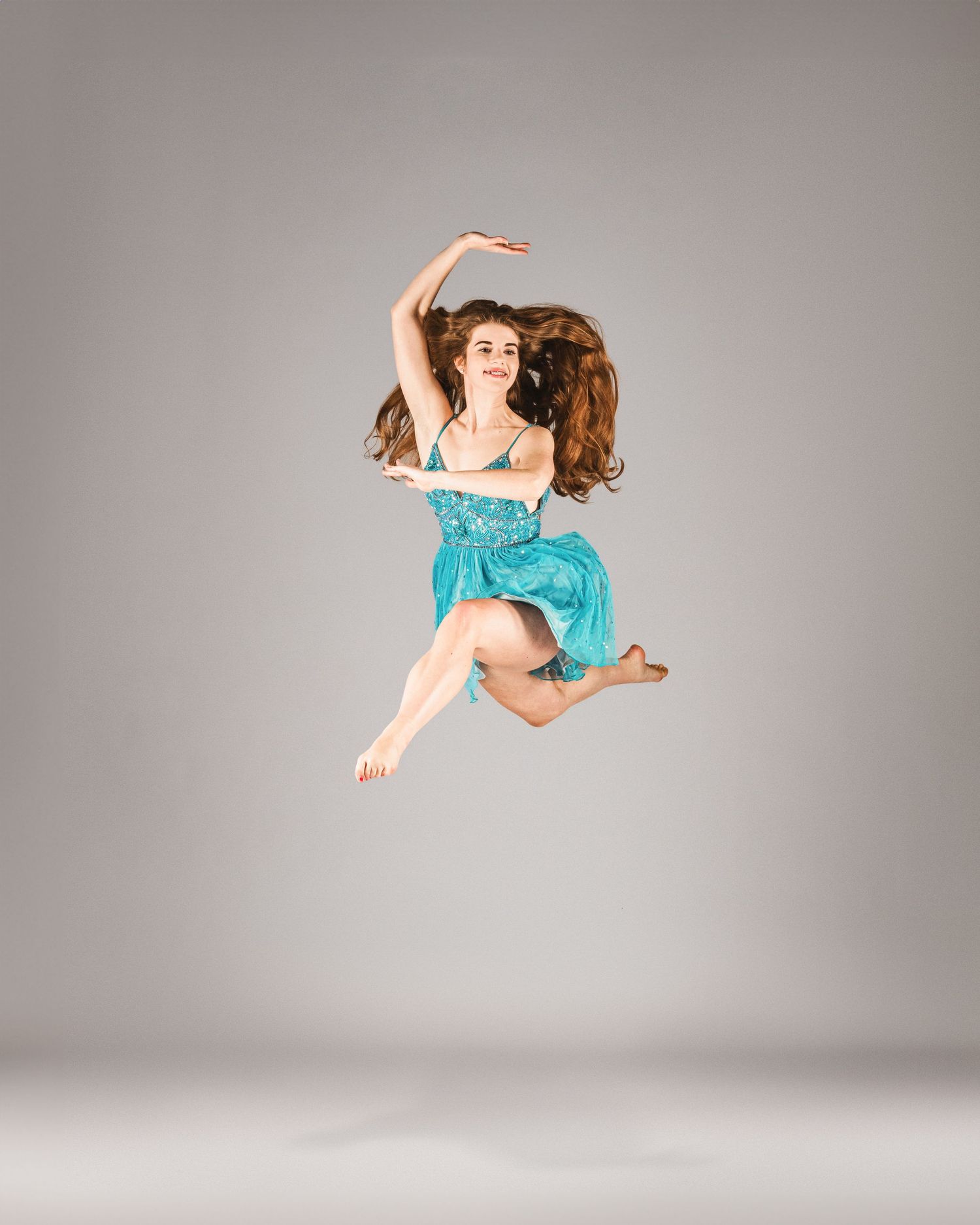 A dancer in a teal dress leaps gracefully mid-air against a gray studio background.