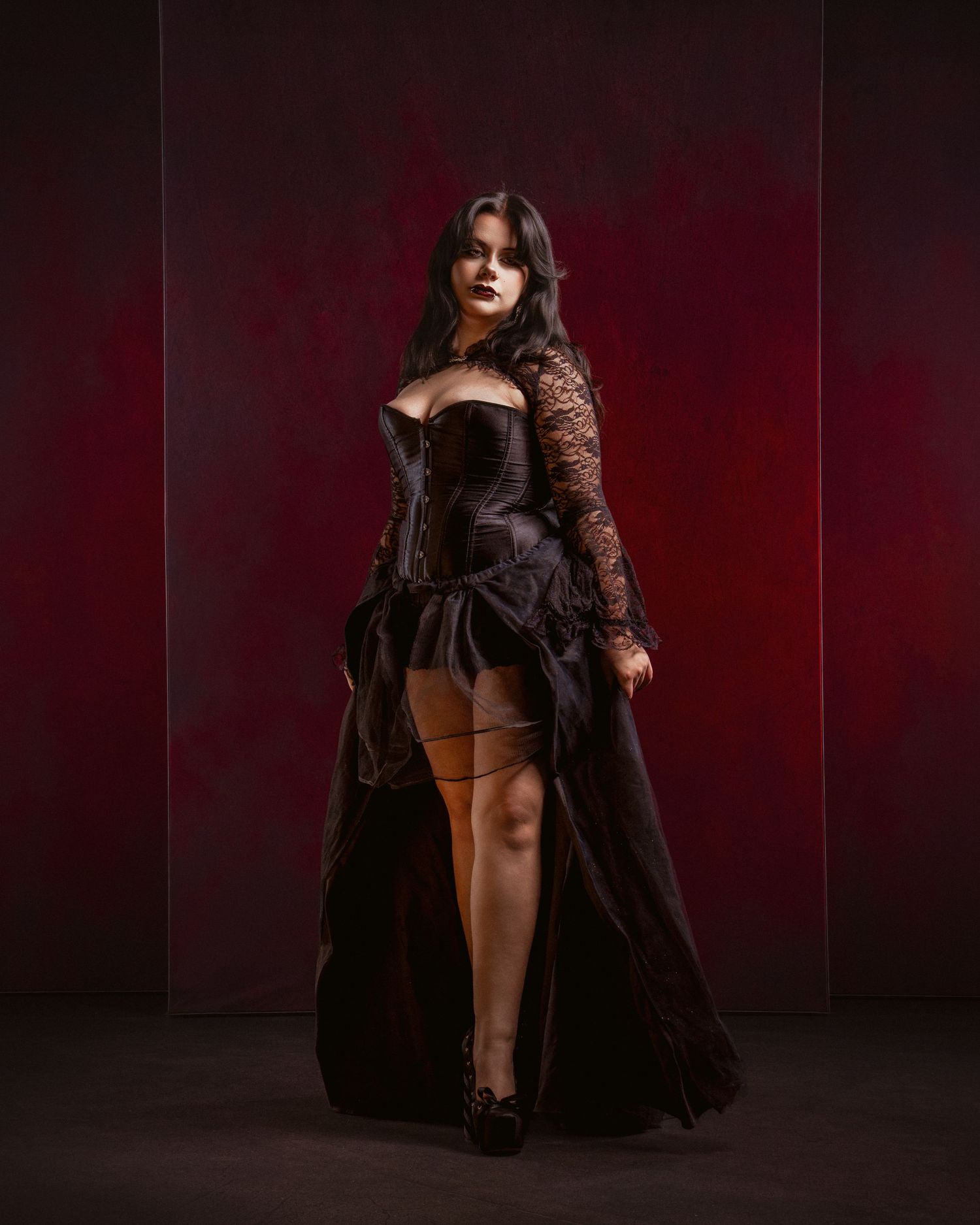 Woman in gothic black corset gown with lace sleeves poses dramatically against deep red background.