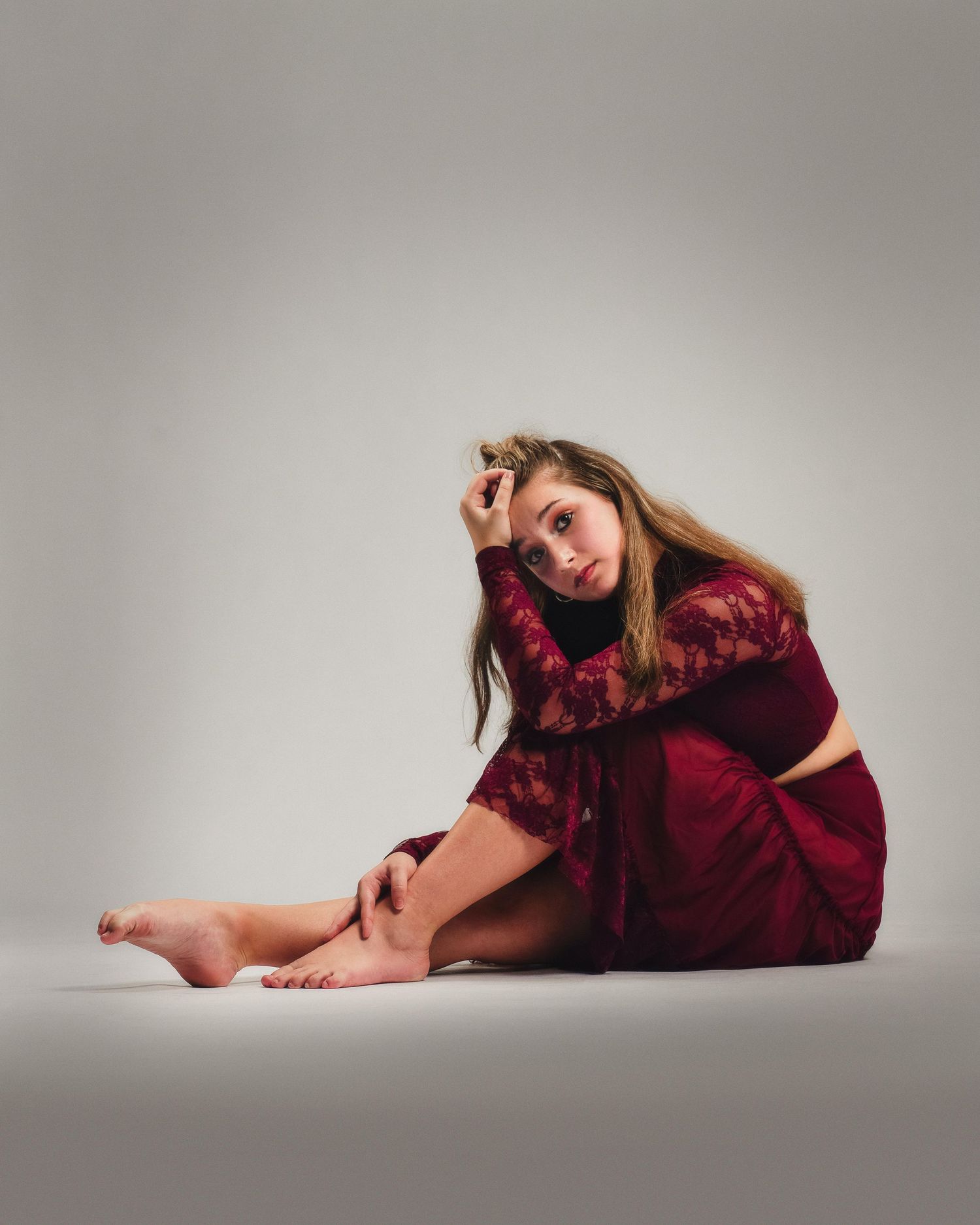 Young woman in burgundy lace outfit seated on floor, posing thoughtfully with hand in hair against gray background.