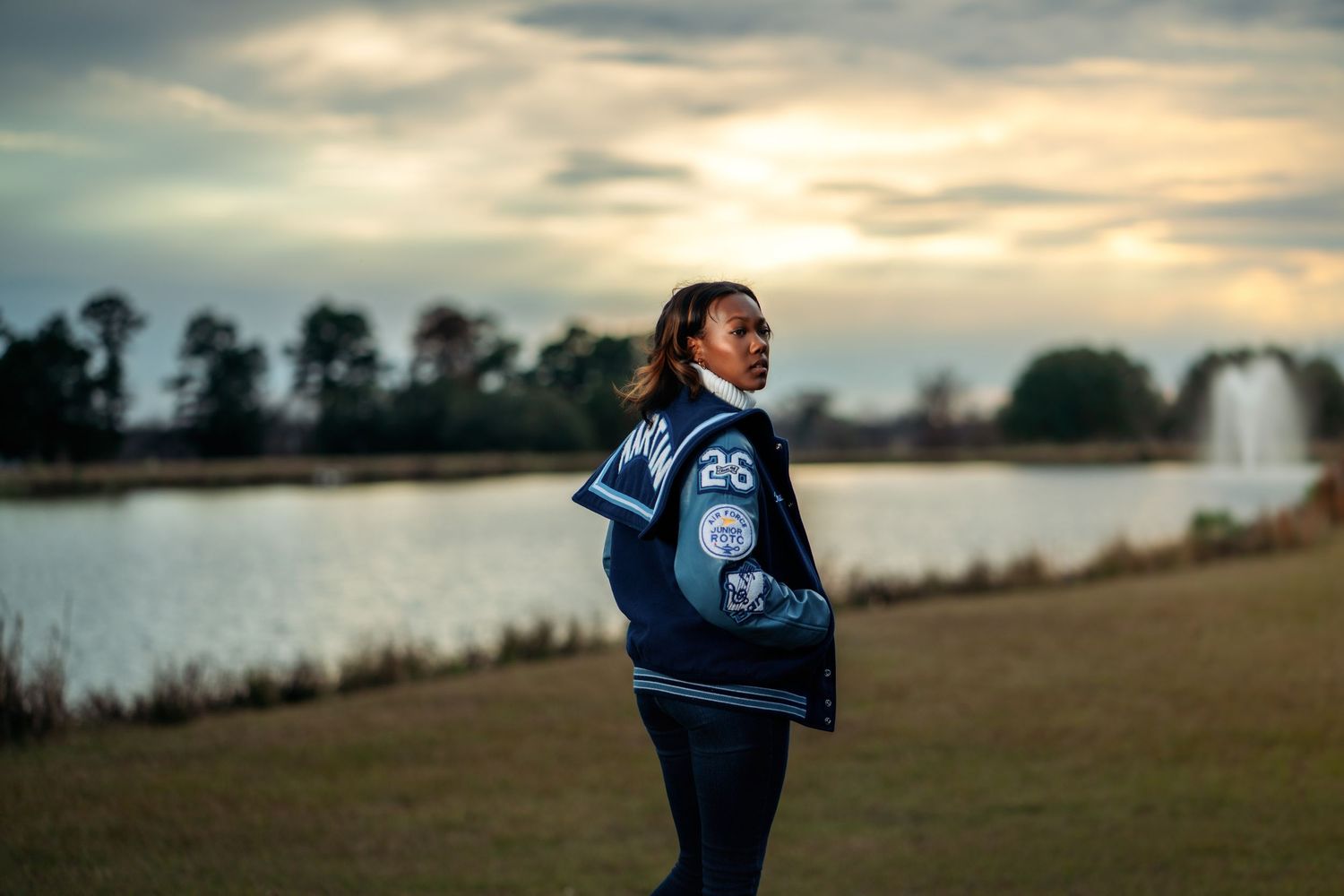 Young woman in varsity jacket number 26 stands by a lake at sunset with fountain and dramatic cloudy sky.