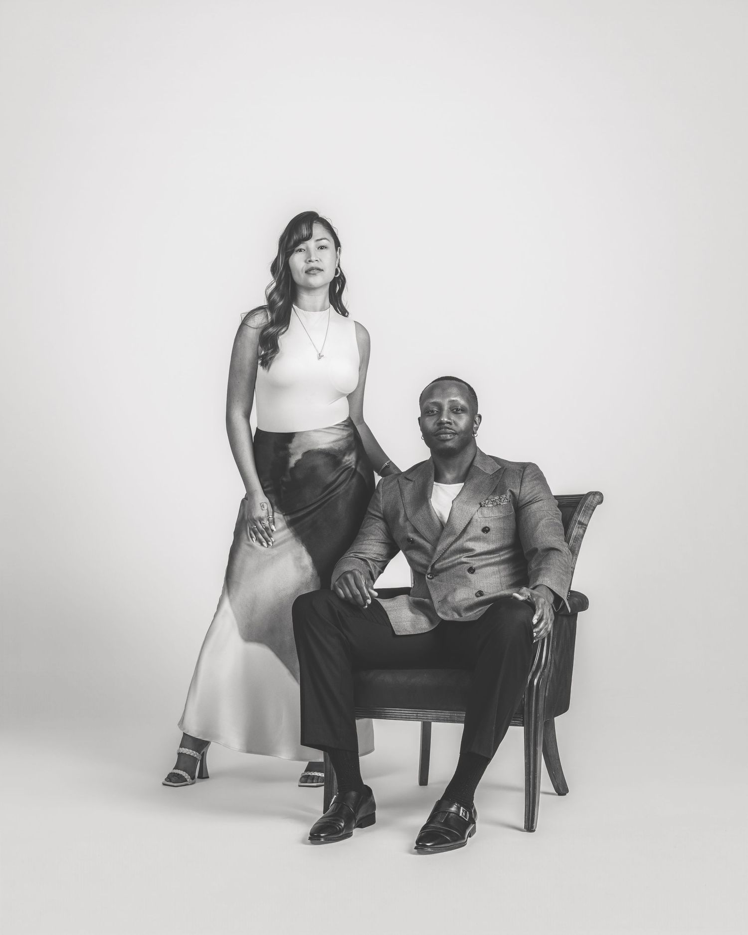 A black and white portrait of a couple posing elegantly, the man seated in a chair, the woman standing beside him.
