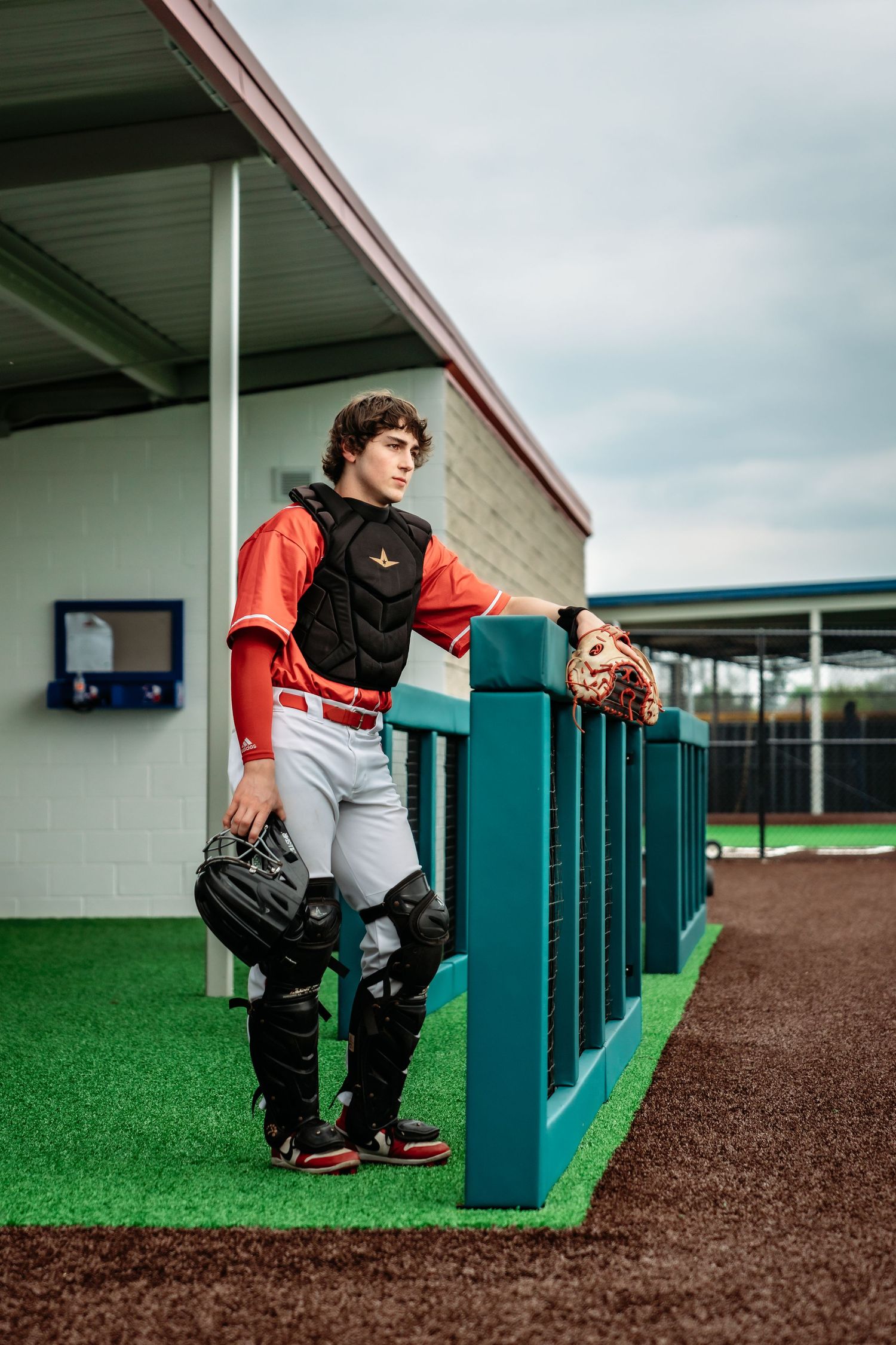 Baseball catcher in full gear leaning on dugout fence at baseball field.