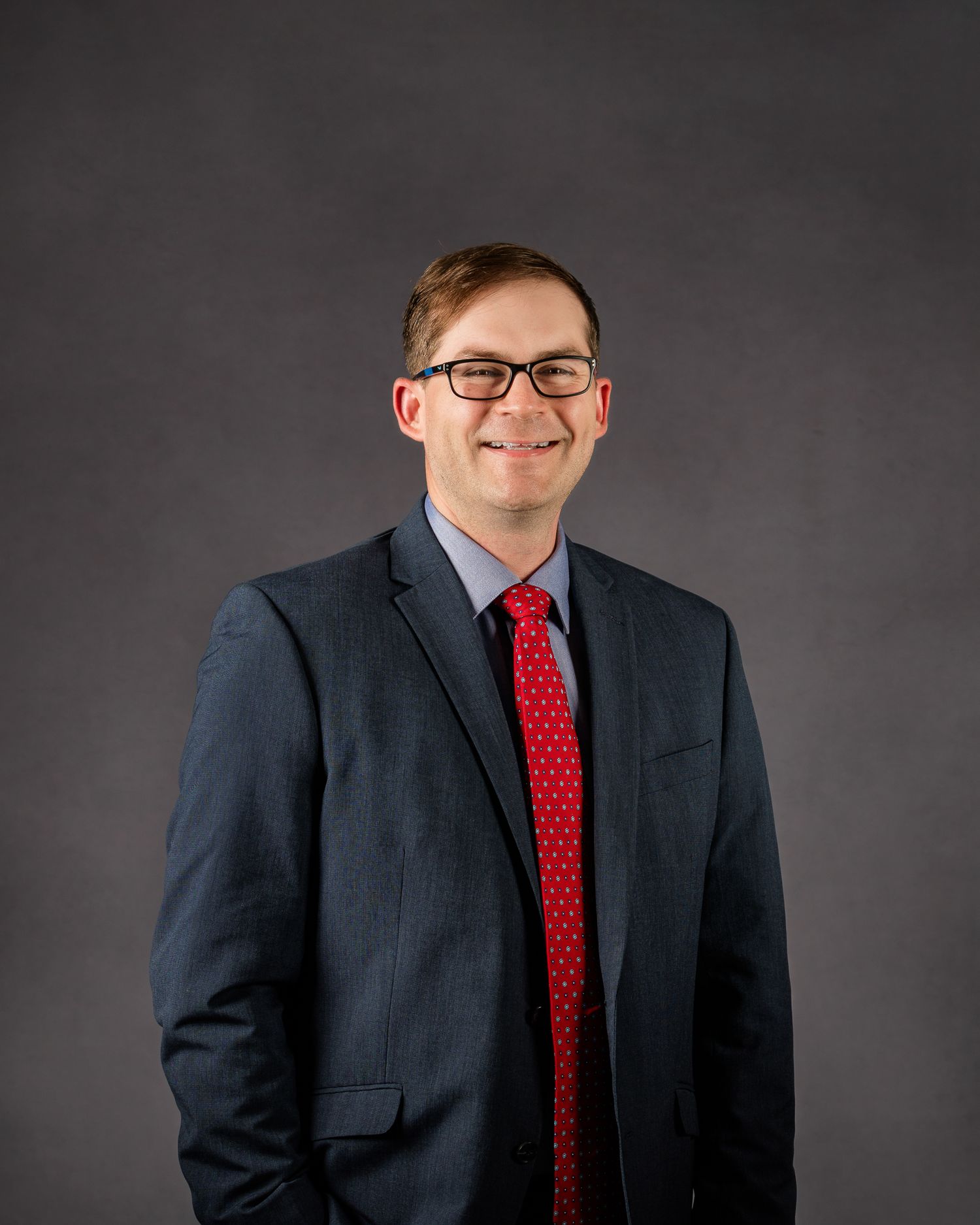 Professional headshot of young man with glasses wearing navy suit and red tie.