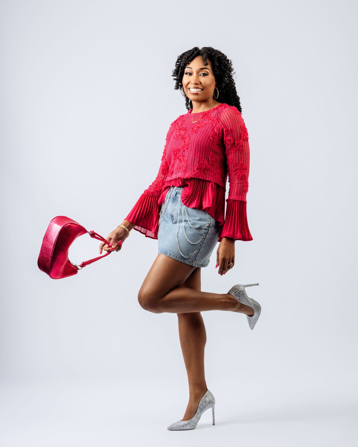 Joyful woman in red sweater and denim skirt posing with heels in studio portrait.