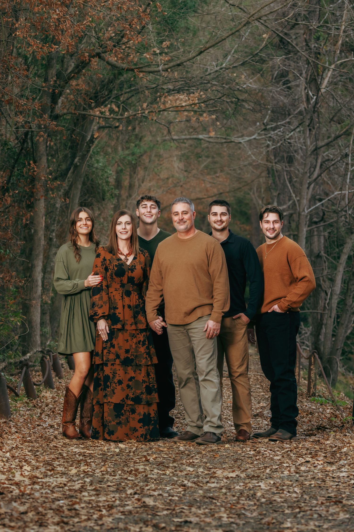 Family of six posing together on a leaf-covered forest path during autumn, wearing warm earth-toned outfits.