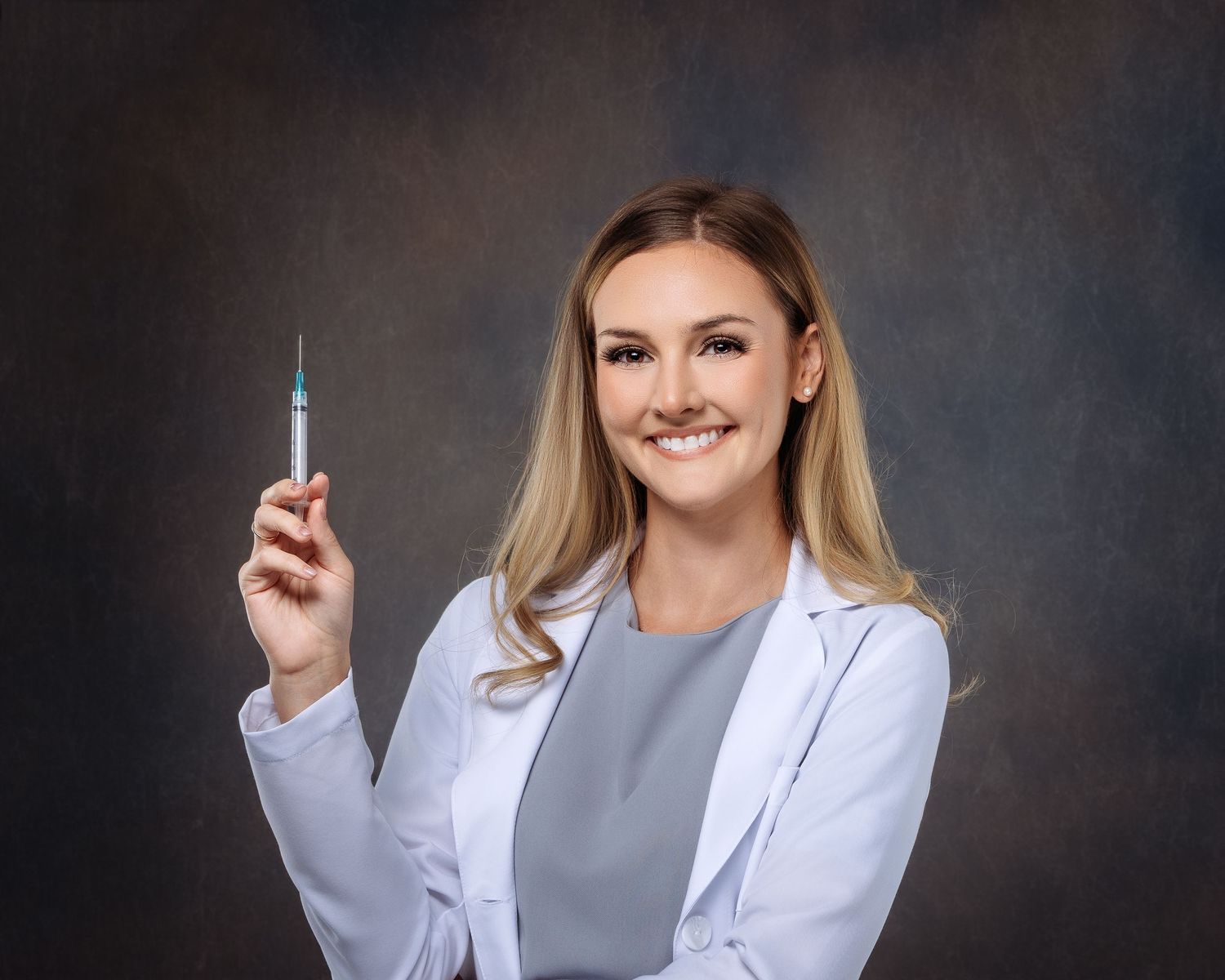 Female healthcare professional in white lab coat smiling while holding a medical syringe against dark background.