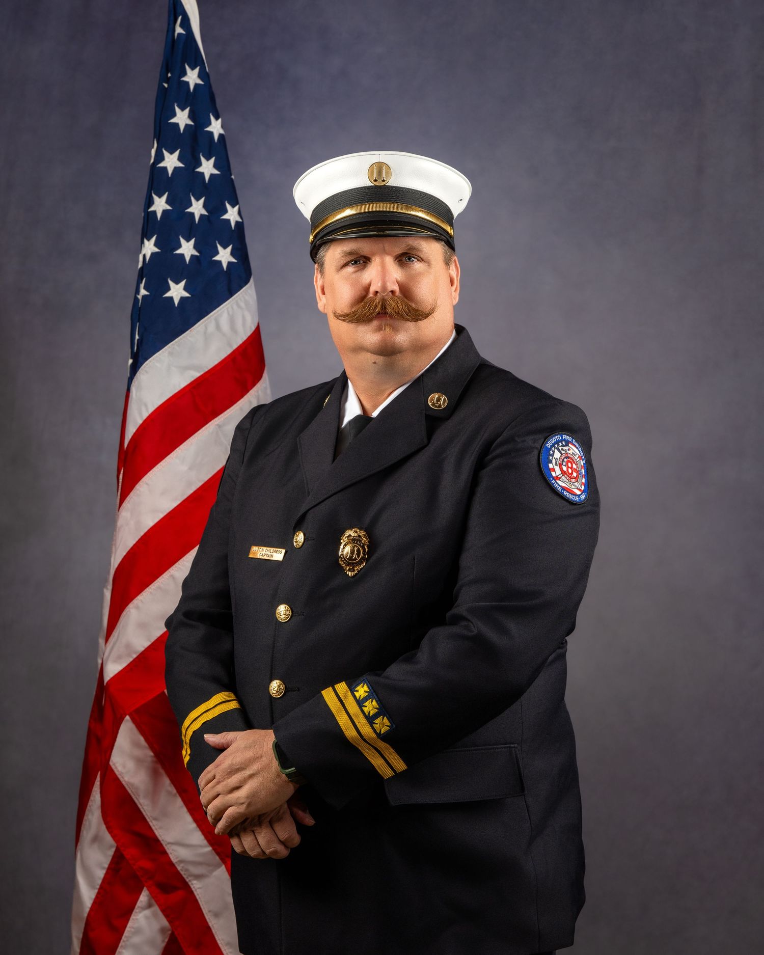 Fire department officer in dress uniform with gold insignia poses beside an American flag for an official portrait.