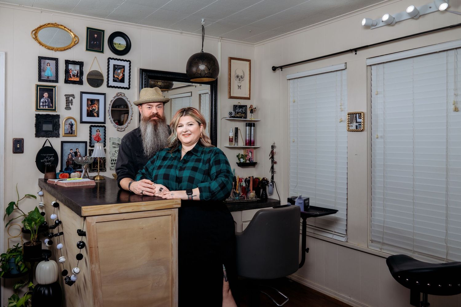 A bearded man and smiling woman pose together in a cozy home salon with gallery wall decor and styling chair.