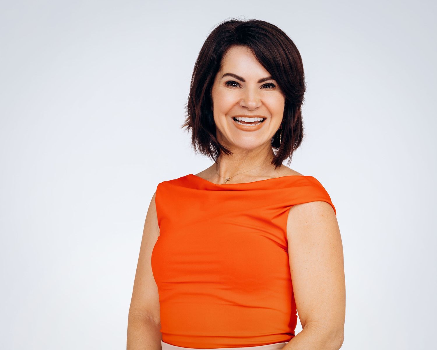 Smiling woman with dark hair wearing an orange sleeveless top, posing against a light gray background.