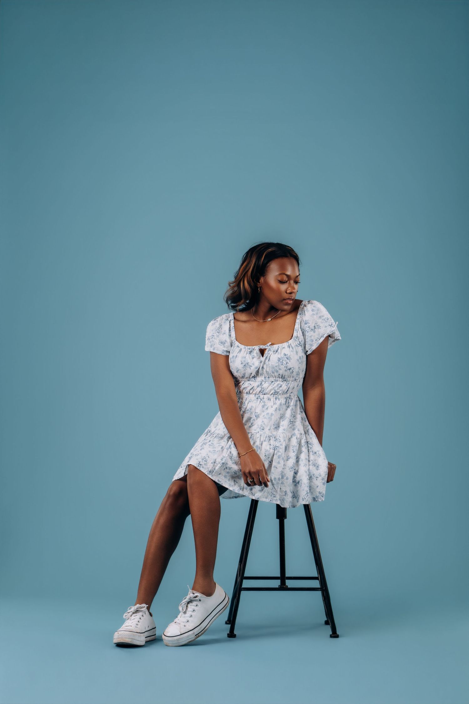 Woman in floral white mini dress sits on stool against blue studio background.