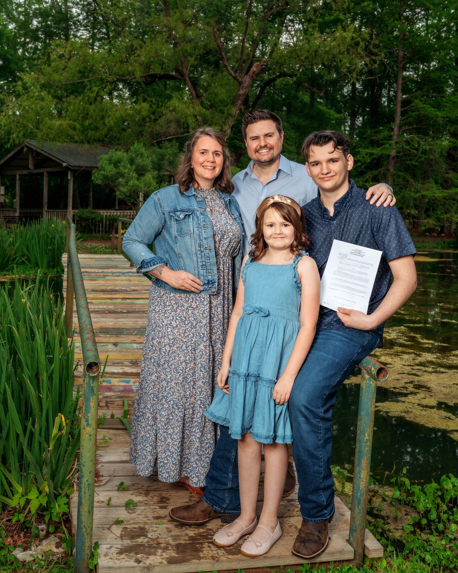 Happy family of four posing on garden bridge holding document near scenic pond outdoors.