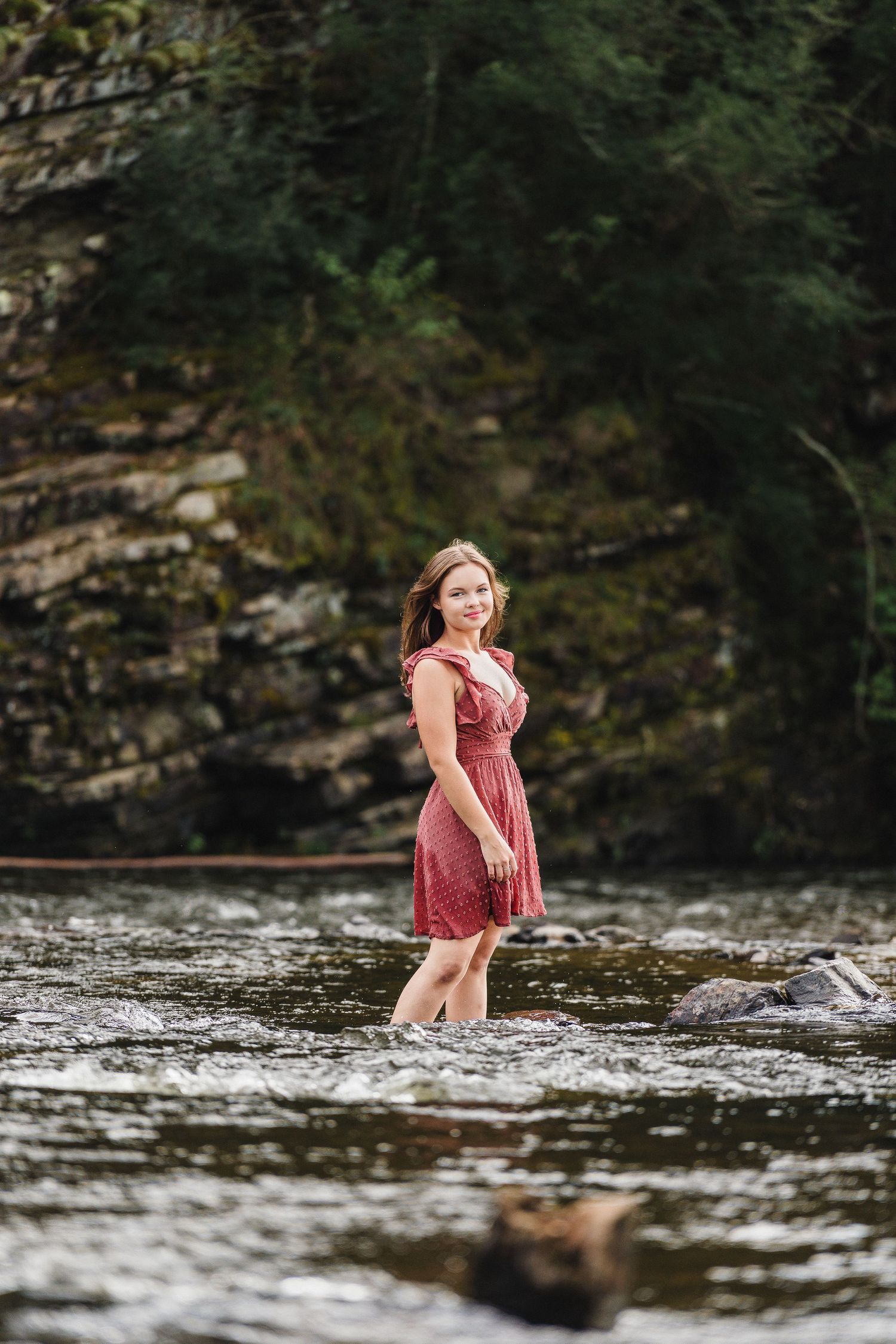 Young woman in red dress wading in a rocky river with stone cliffs behind her.