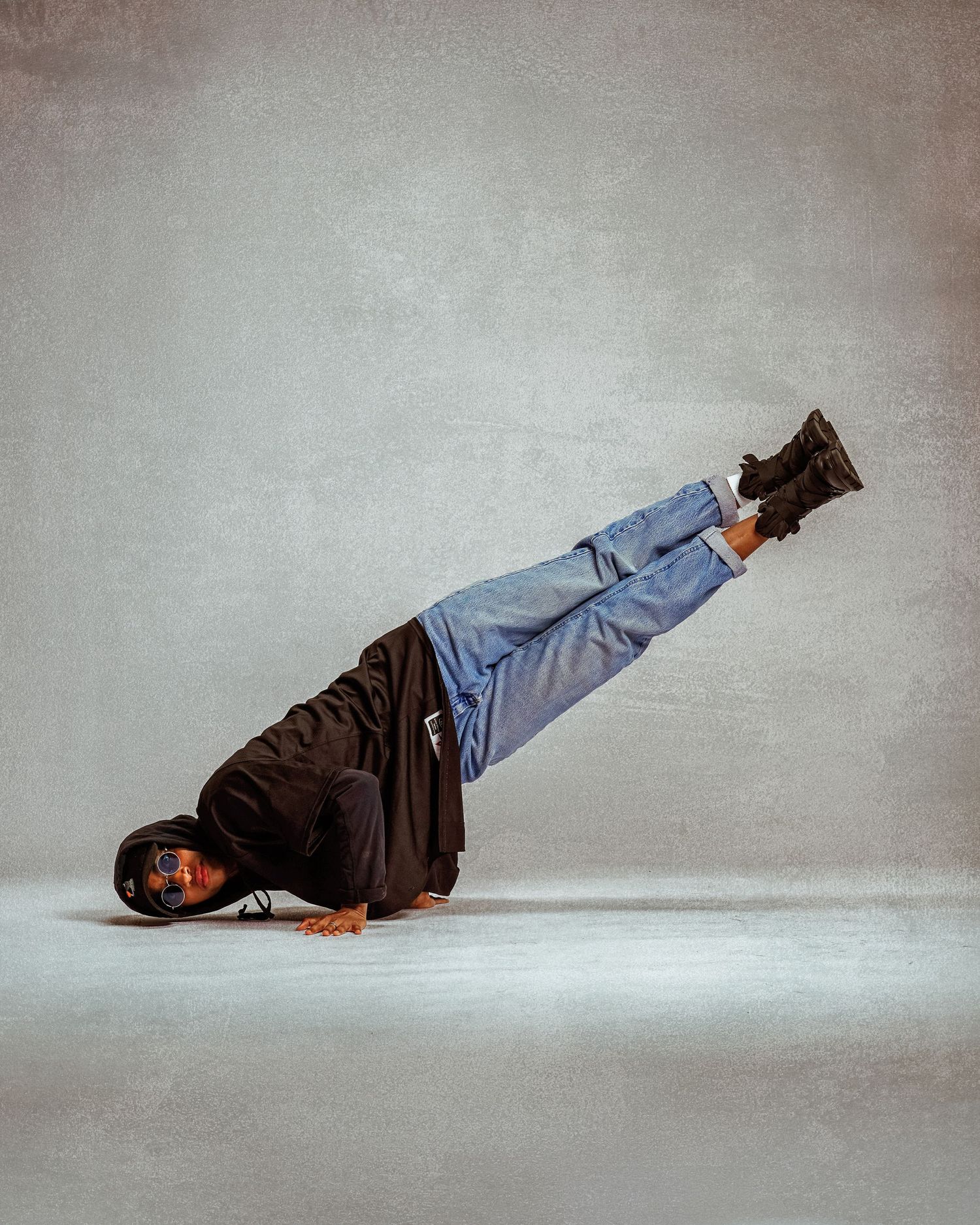 Breakdancer balancing on one hand with legs raised in jeans and black hoodie on studio floor.