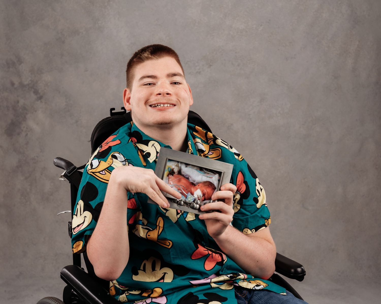 Smiling young man in a Mickey Mouse shirt sitting in a wheelchair, holding a framed photo against a gray background.