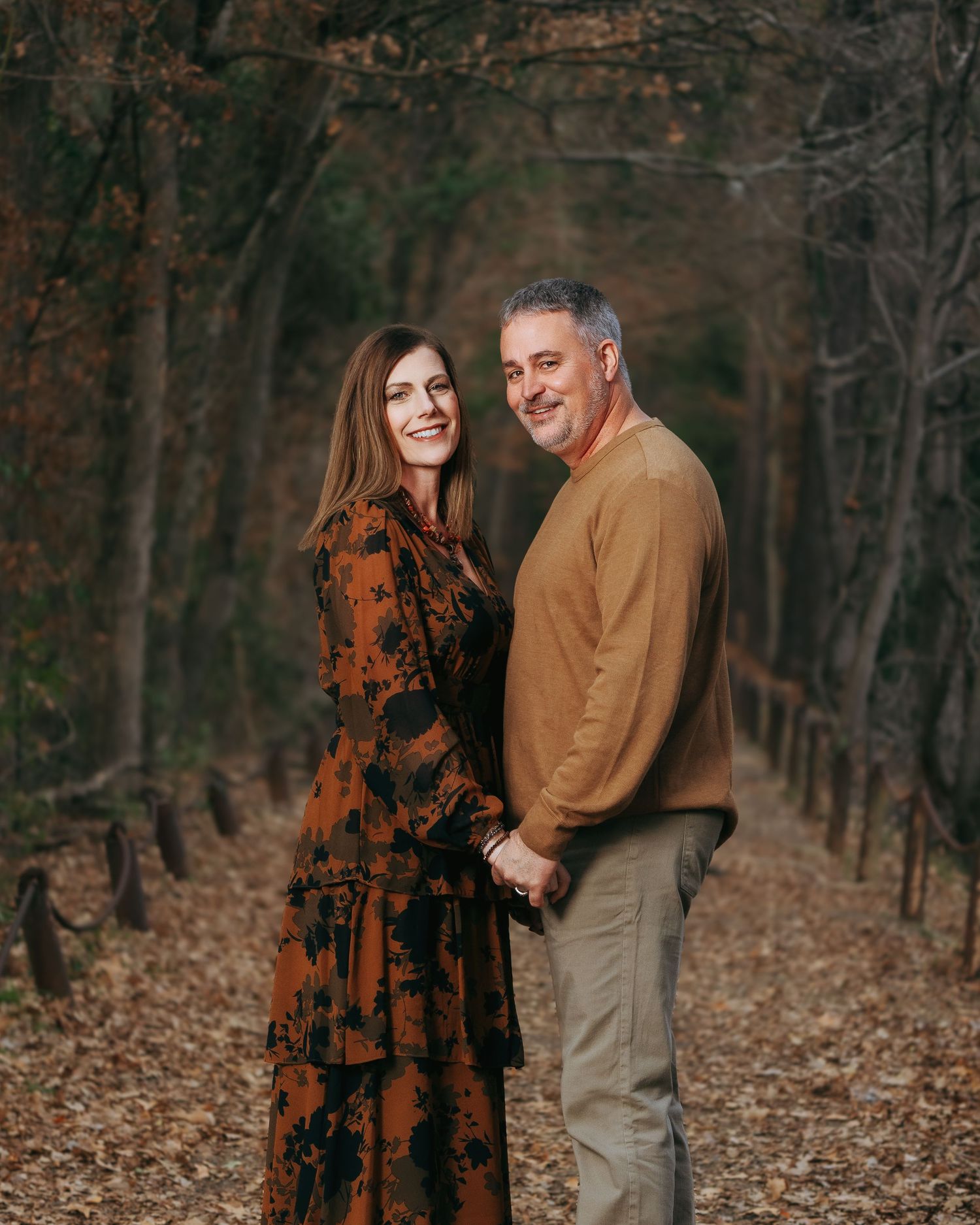 Couple holding hands and smiling on a scenic autumn forest path, woman in floral maxi dress and man in tan sweater.