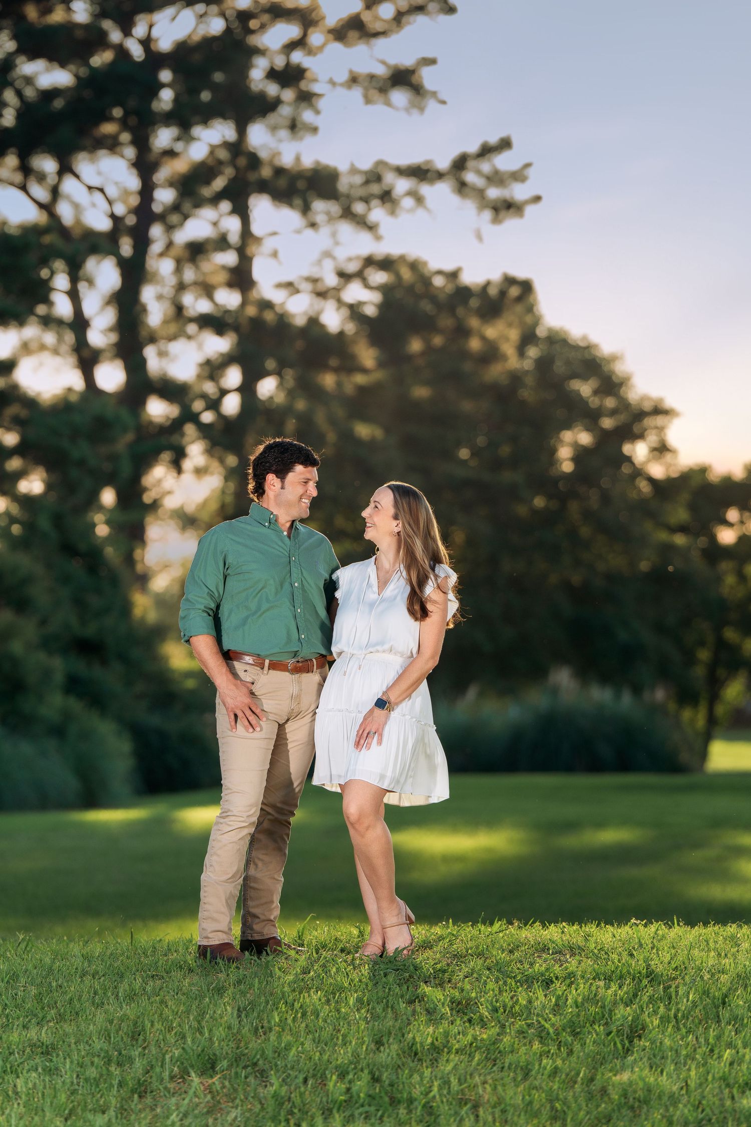 A smiling couple in casual summer attire shares a joyful moment on a green lawn at sunset.
