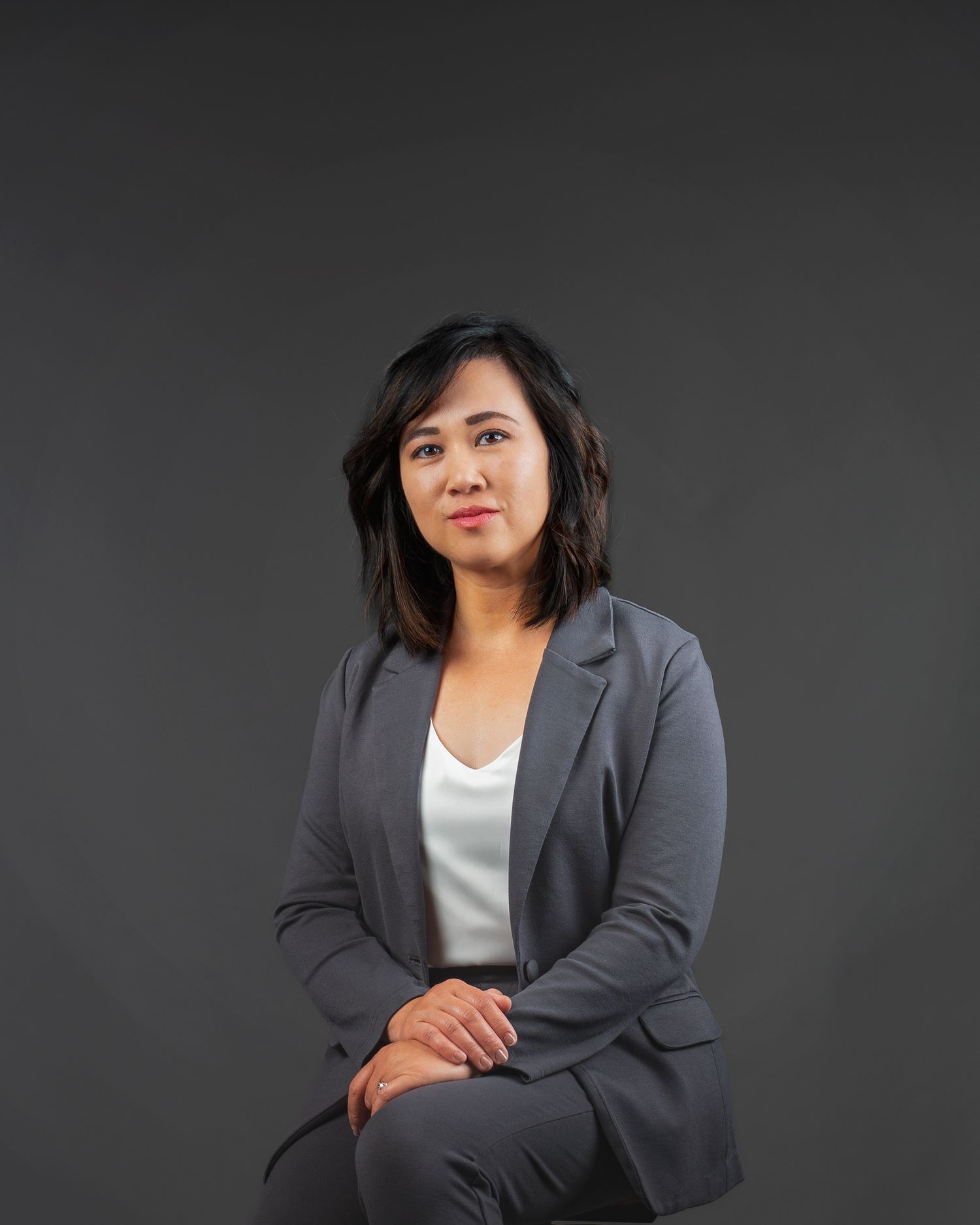 Professional woman in gray blazer sitting confidently against dark background for corporate headshot.