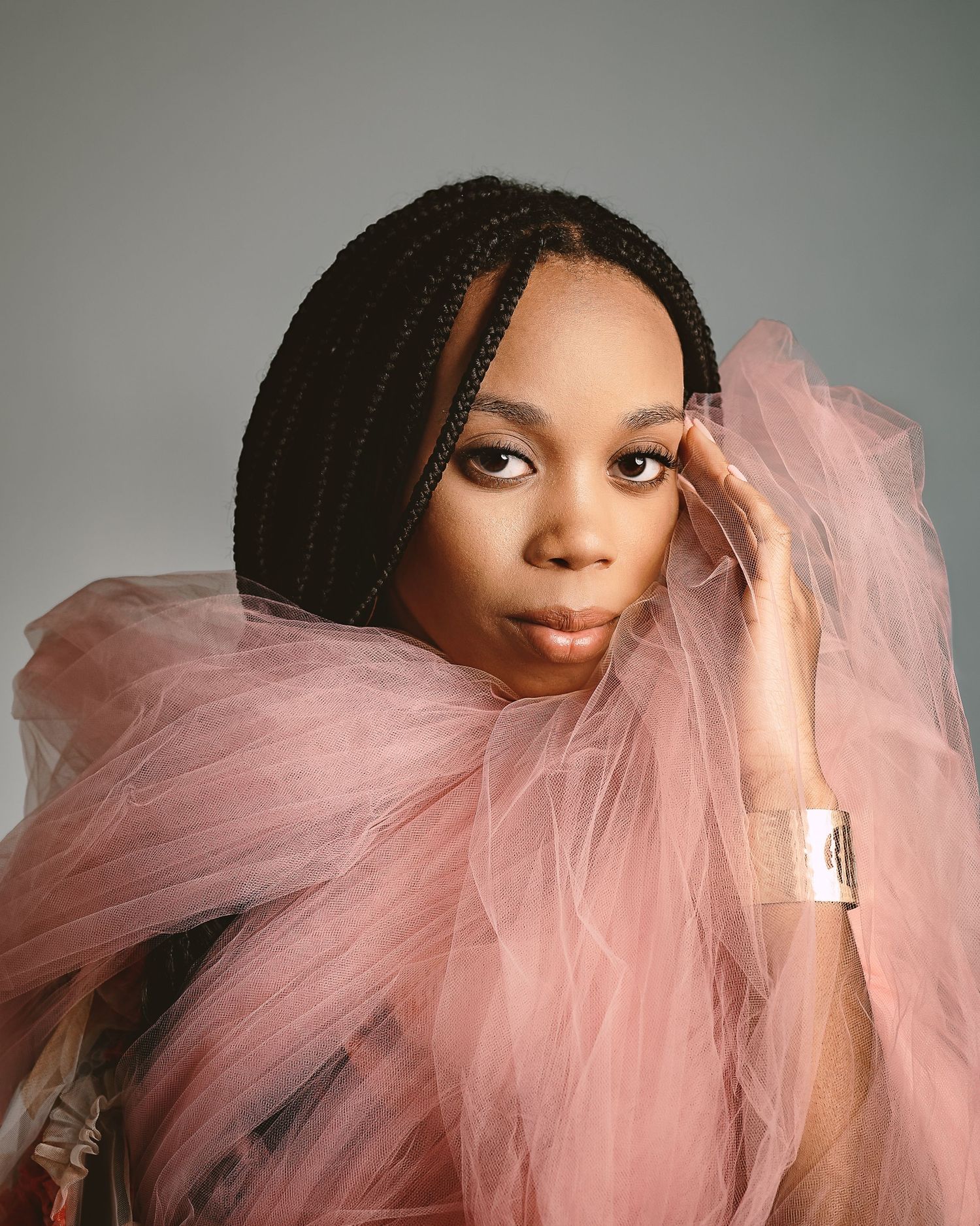 Young Black woman with box braids poses in a dramatic pink tulle garment against a gray backdrop.