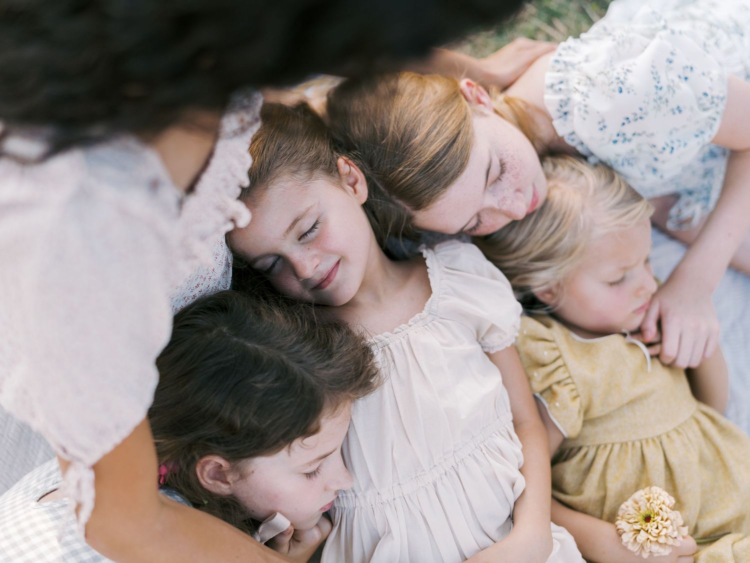 A mother and four young children cuddle together outdoors in soft natural light, wearing pastel floral dresses.