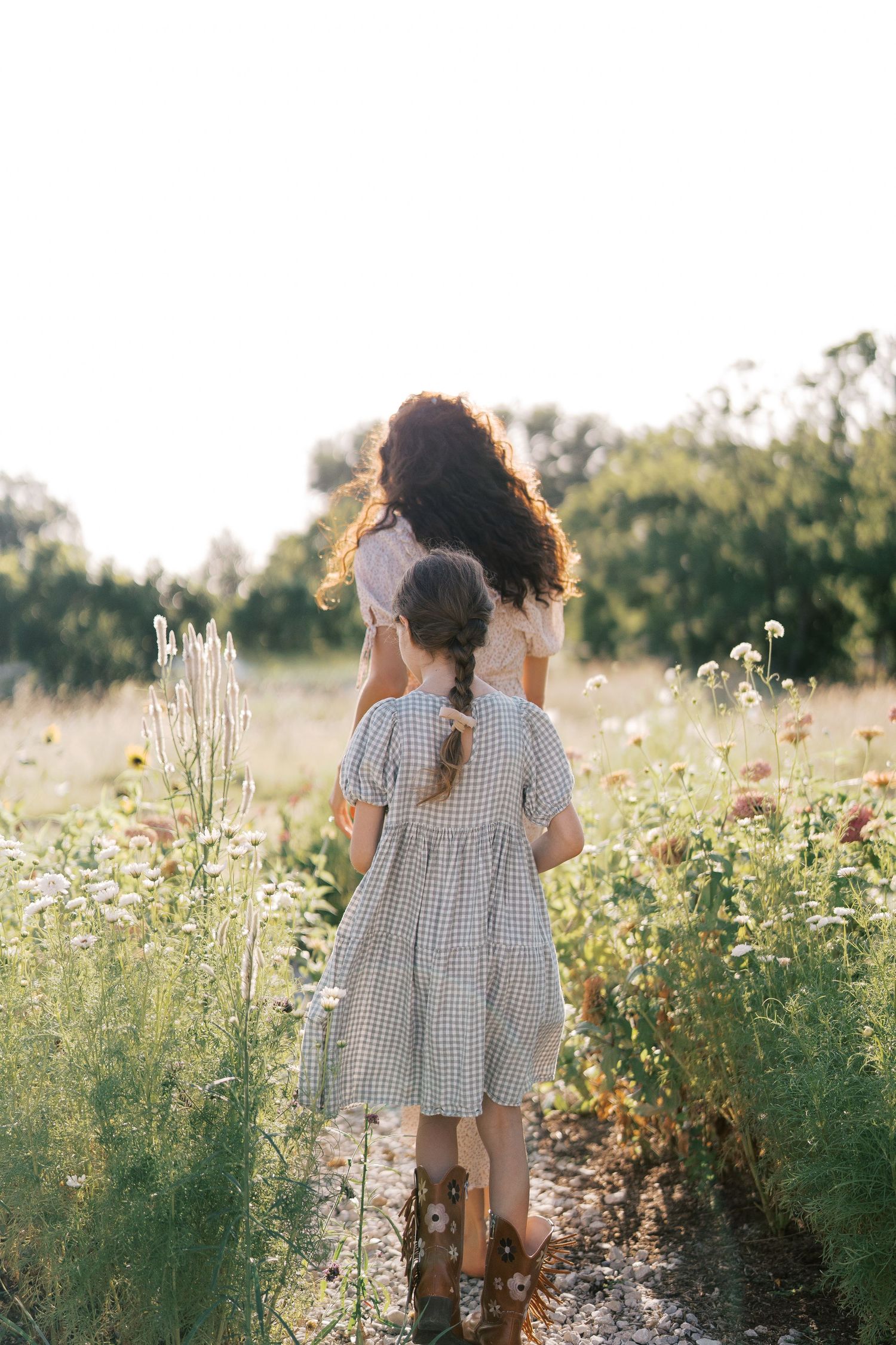 A woman and young girl in dresses walk through a wildflower meadow path, viewed from behind in golden sunlight.