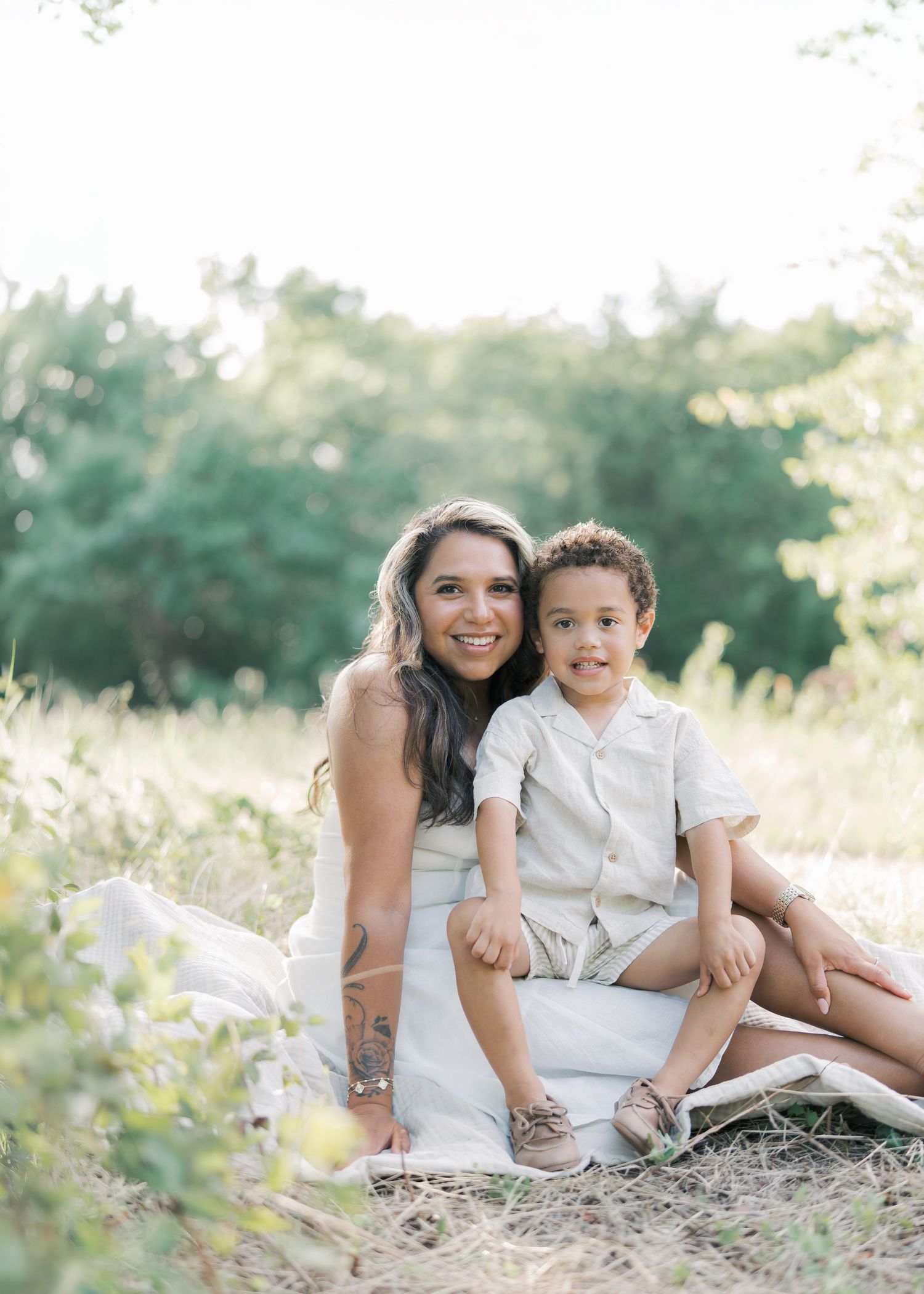 A smiling mother sits outdoors in a field with her young son in a beige outfit on her lap by Alahir Villasenor Photographer