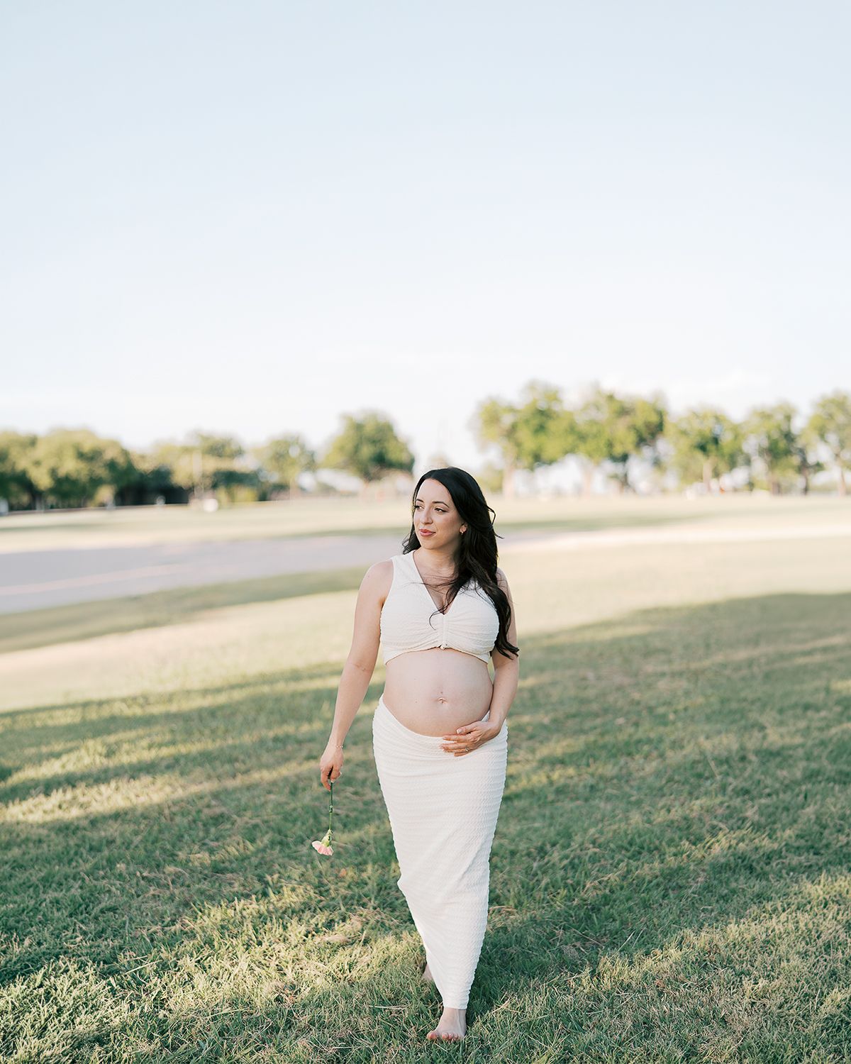 Pregnant woman in white crop top and skirt cradling baby bump while walking barefoot in a sunny green park.
