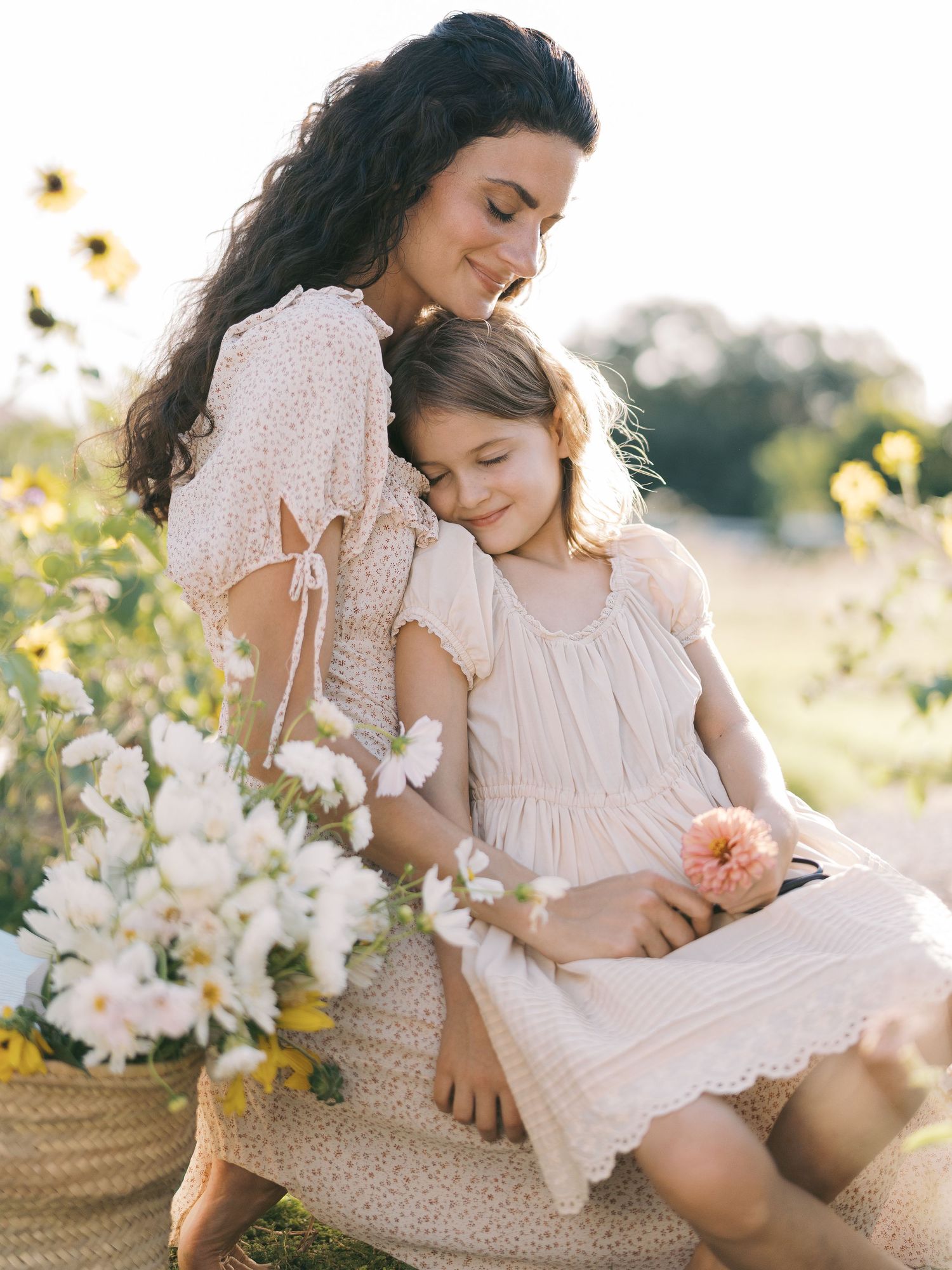 A mother tenderly embraces her young daughter outdoors, surrounded by white and yellow flowers in warm sunlight.