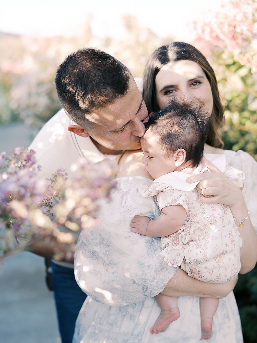 Loving parents lean in to kiss their newborn baby girl in a floral outfit among purple flowers by Alahir Villasenor Photographer