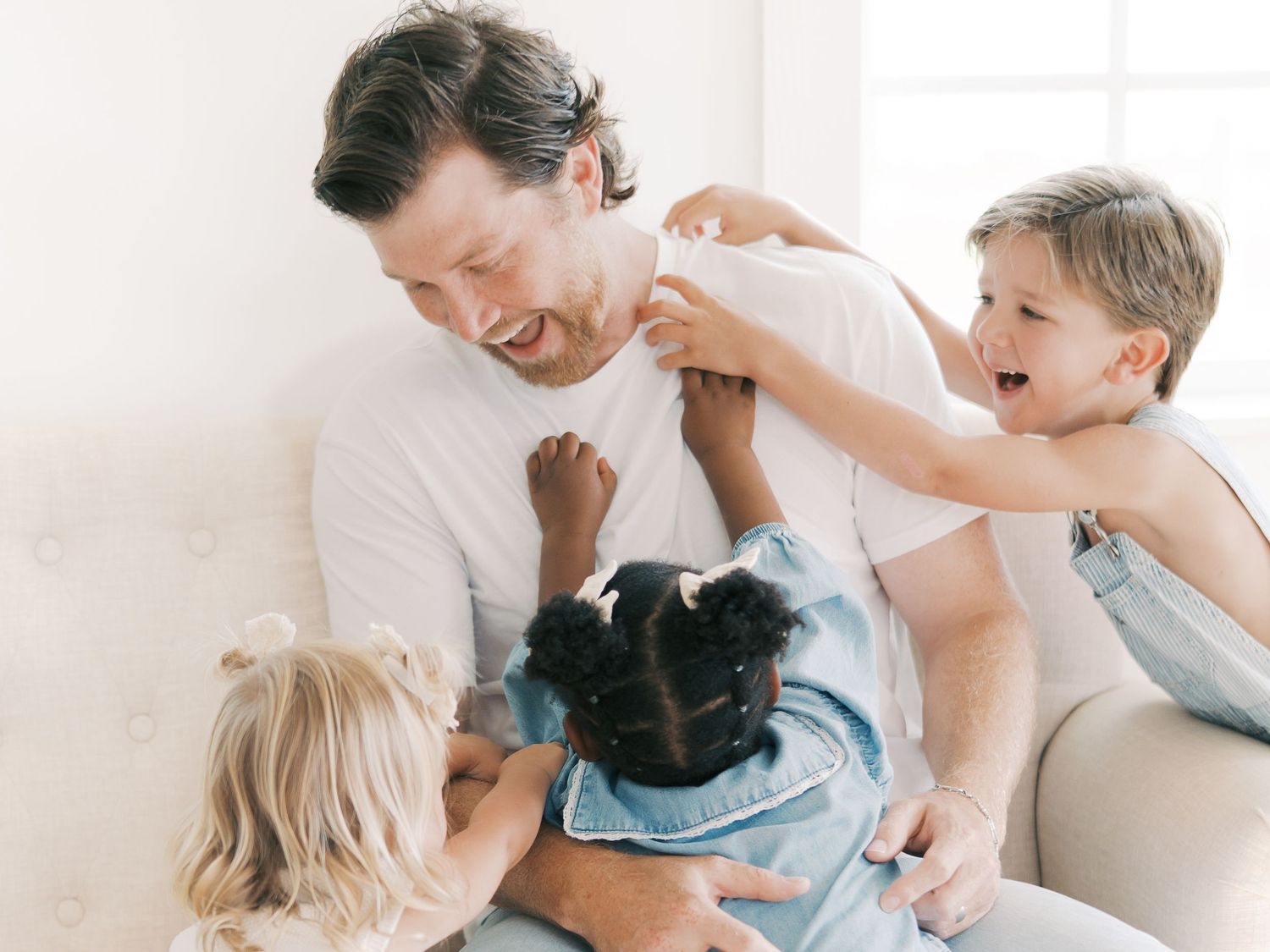 Laughing father plays with three energetic children climbing on him in a bright room in Dallas by Alahir Villasenor Photography-FamilySessio