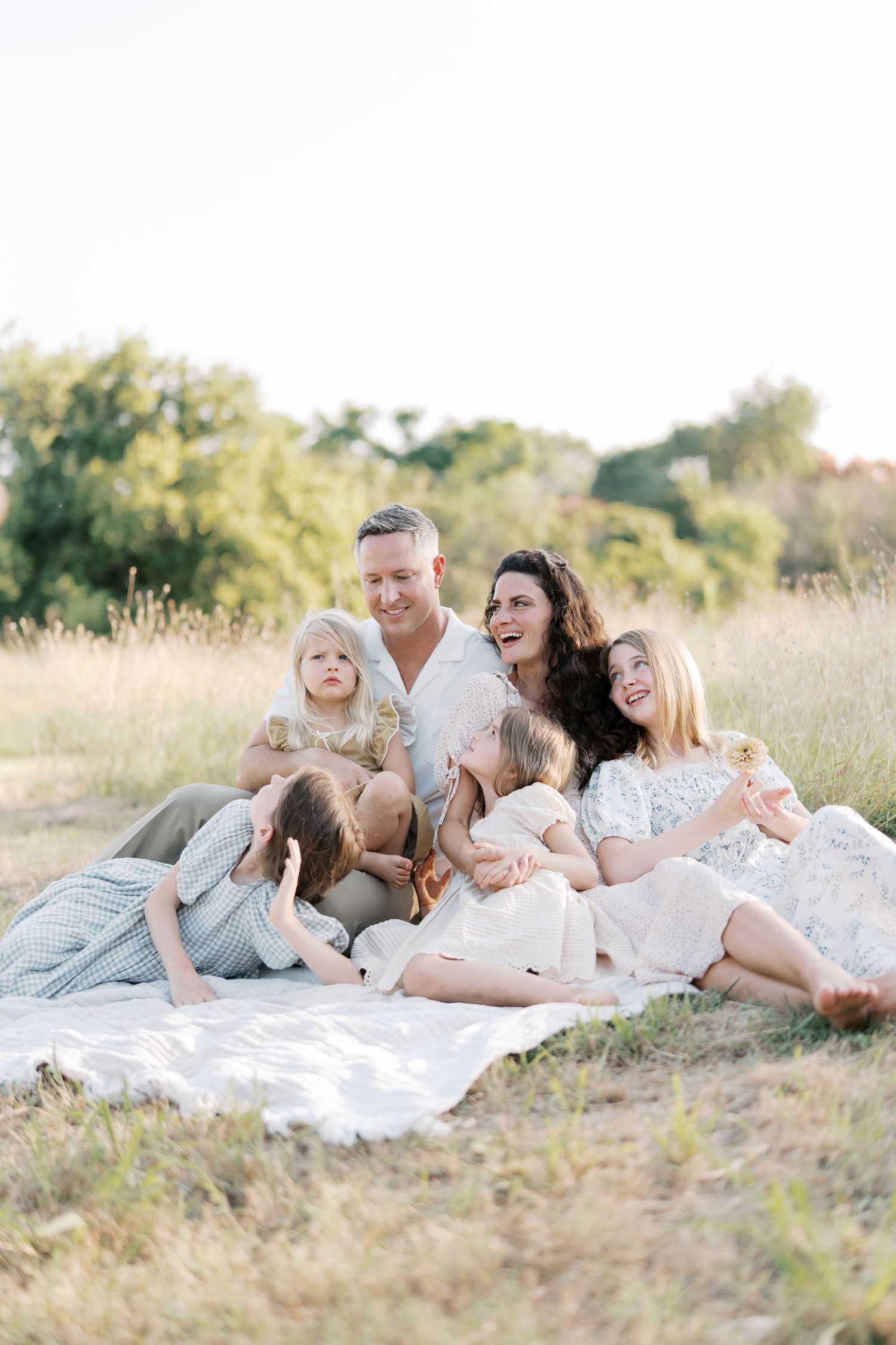 A joyful family of six sits together on a blanket in a golden sunlit meadow, laughing and playing.