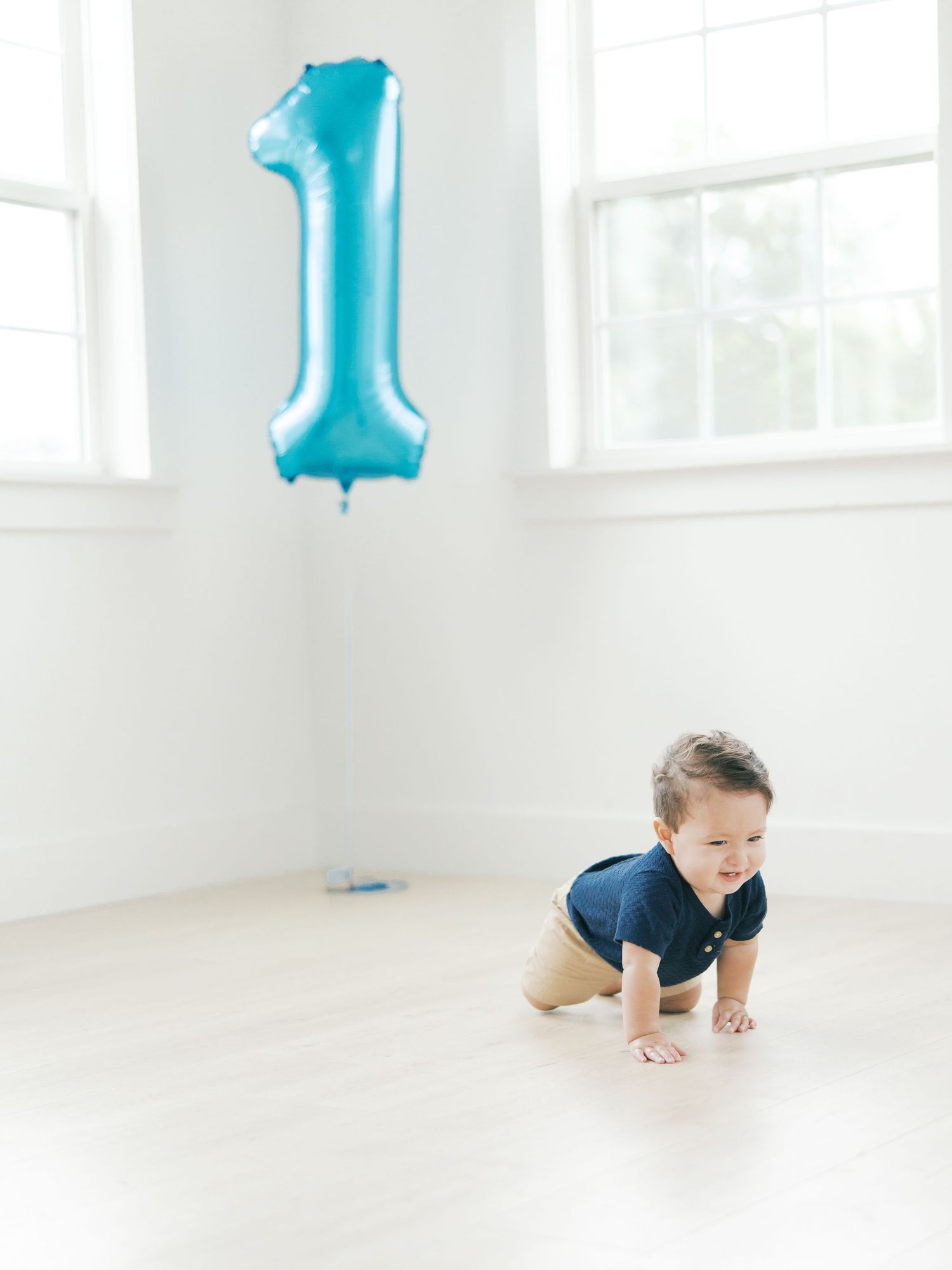 A baby boy crawls on a bright white floor near a blue number one balloon celebrating his first birthday.