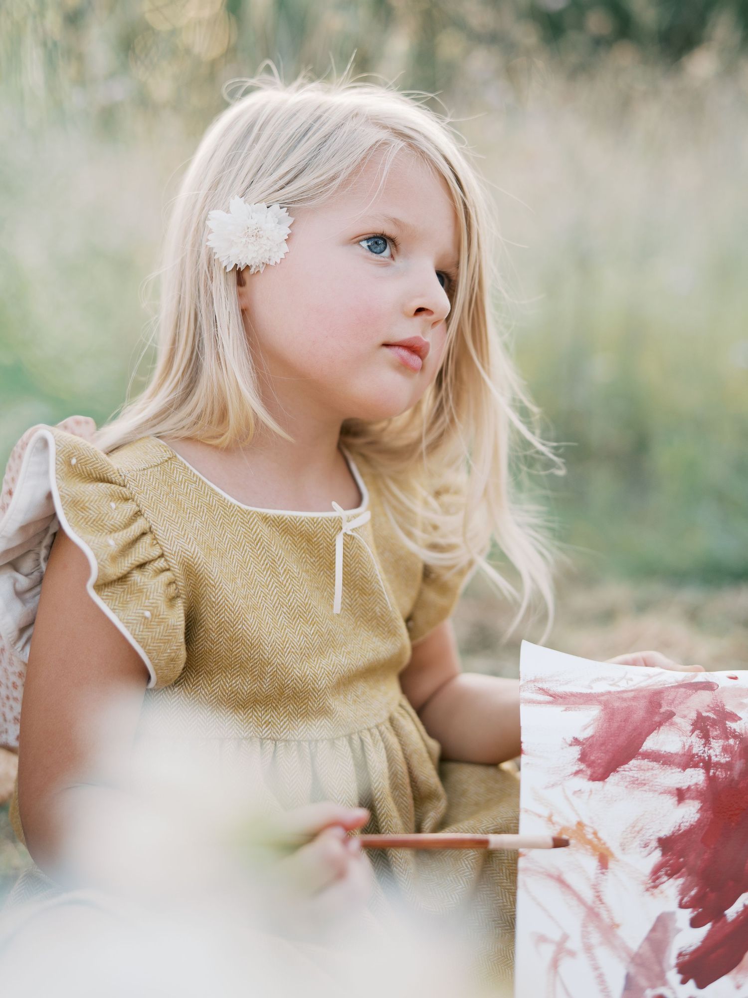 A young blonde girl with a white flower hair clip wears a golden dress outdoors in a softly blurred natural setting.