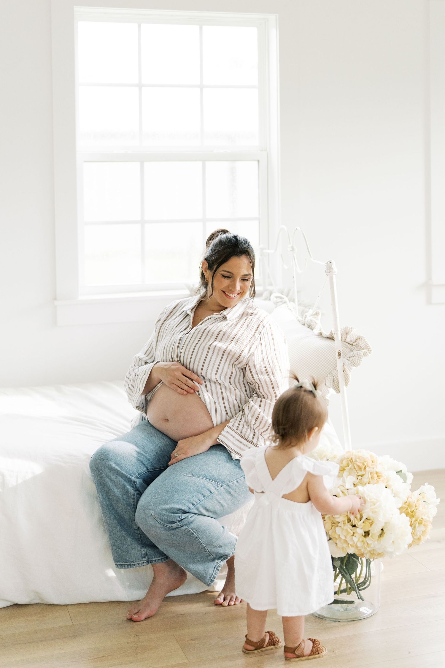 Pregnant woman in striped shirt and jeans sitting with toddler girl near white flowers.