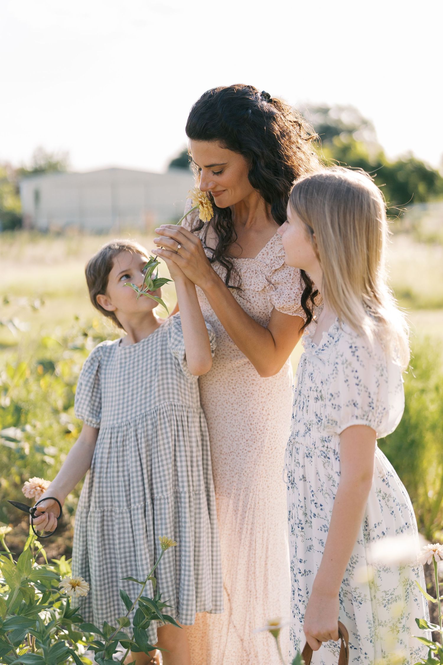 A mother and two daughters smell wildflowers in a sunlit field, wearing soft floral and gingham dresses.