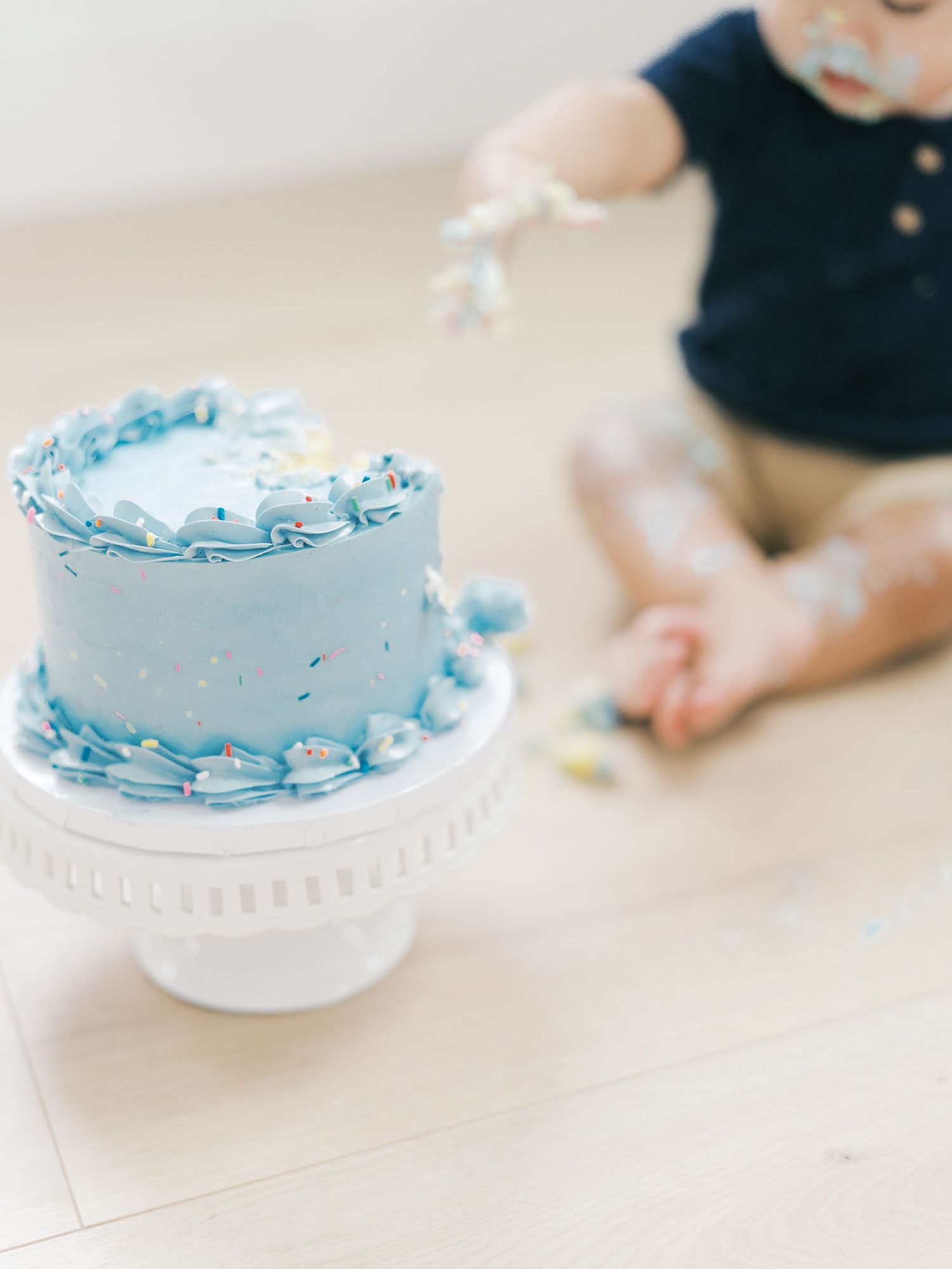 Blue confetti smash cake on white stand with baby boy in background during birthday session.