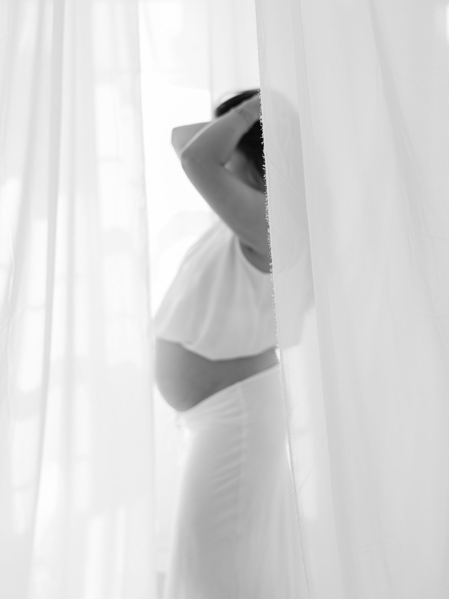 Black and white maternity photo of pregnant woman in white dress standing behind sheer curtains in Dallas by Alahir Villasenor Photography.