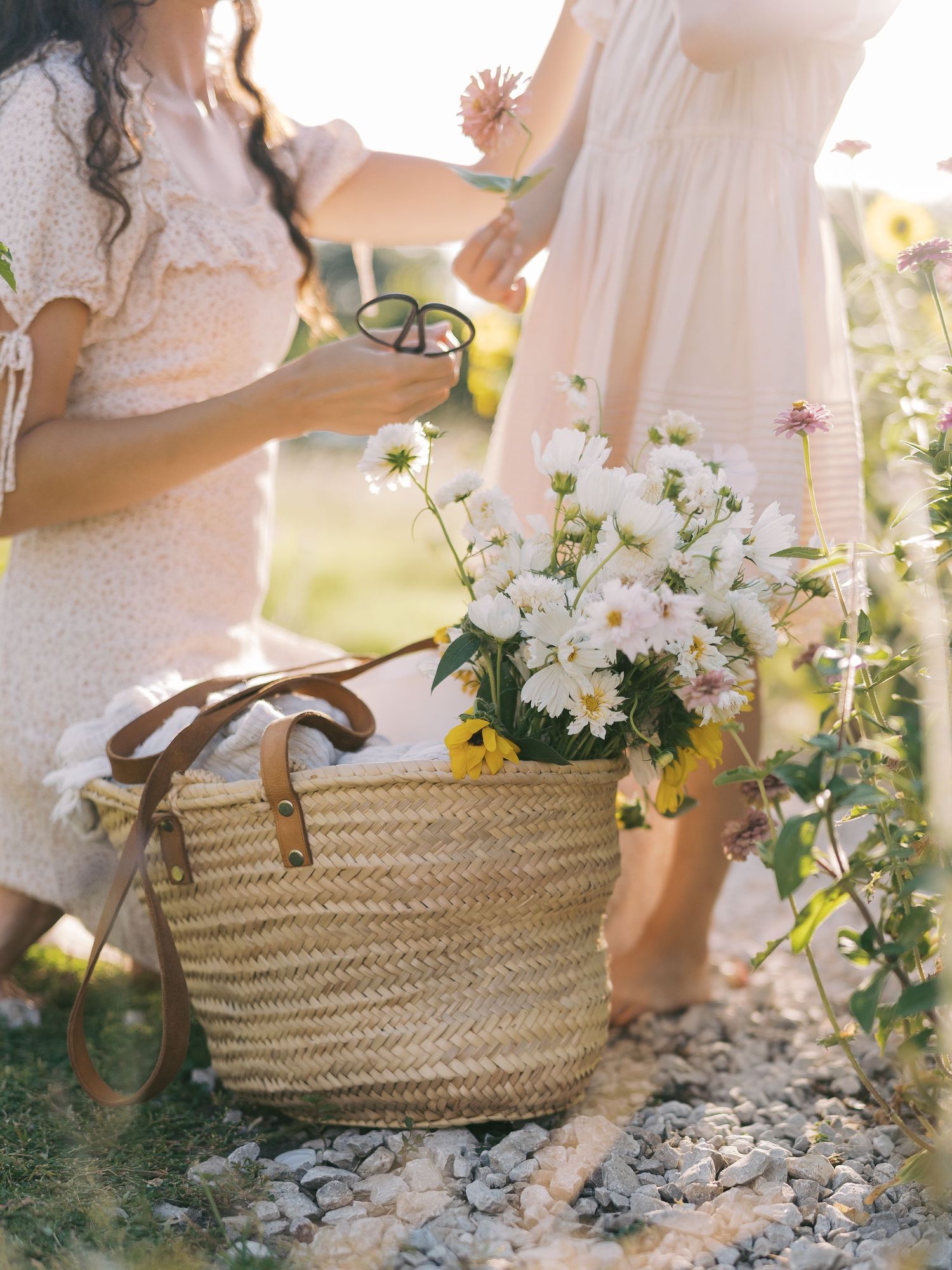 Two women in white dresses arranging fresh white flowers in a woven straw basket outdoors.