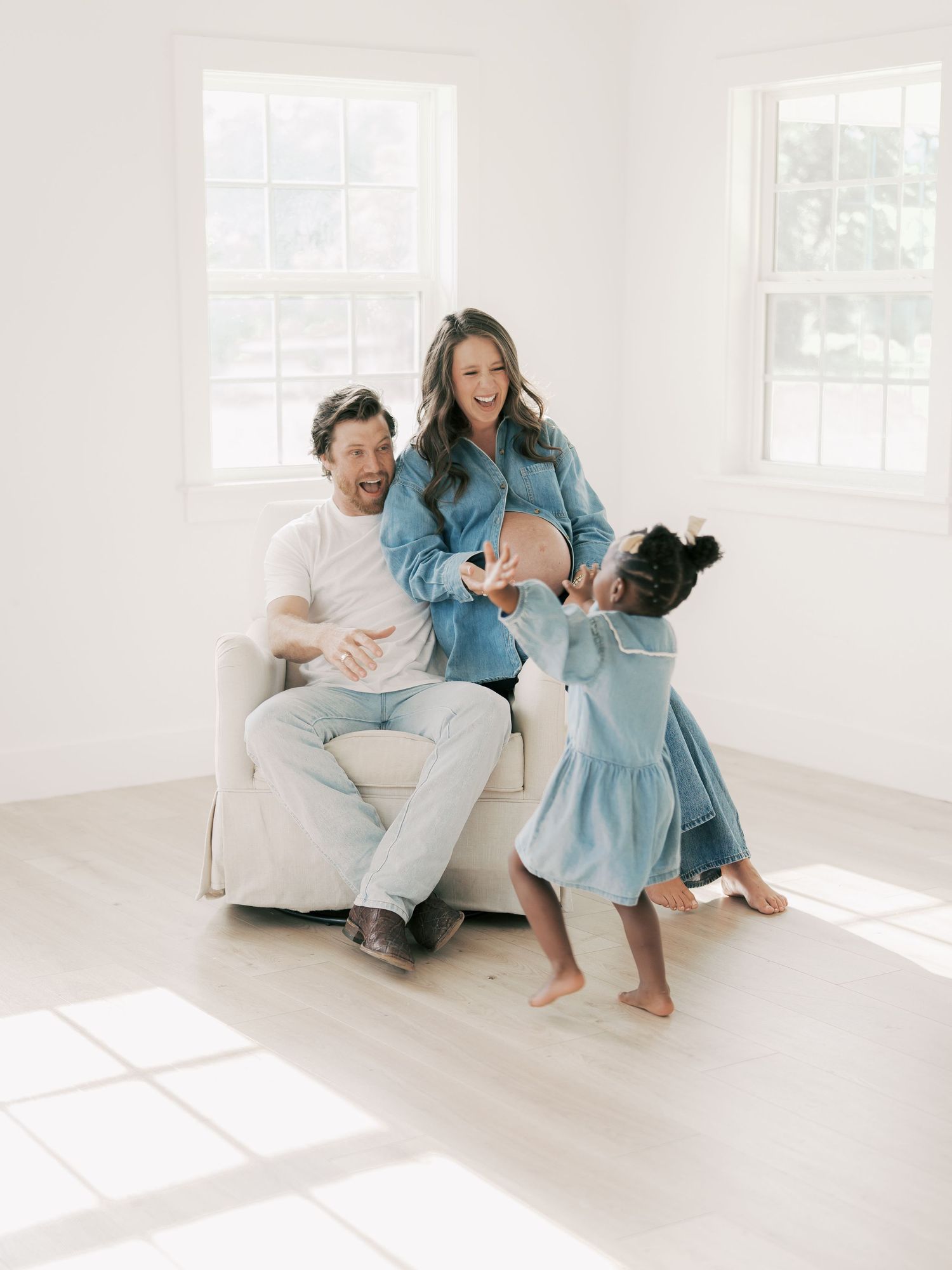 Smiling pregnant couple in denim sit with their excited young daughter in bright studio Dallas by Alahir Villasenor Photography-FamilySessio