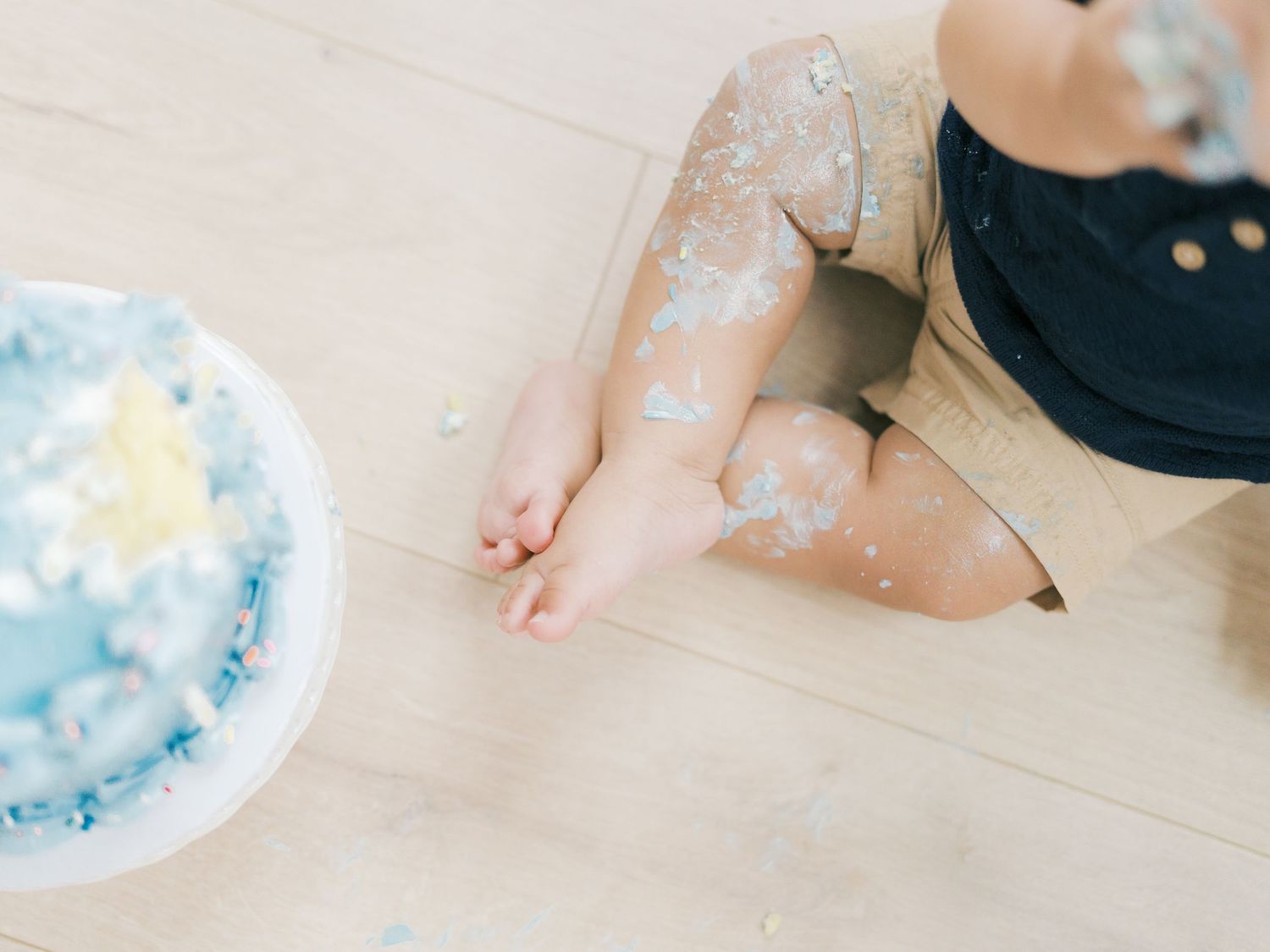 Close-up of baby legs covered in frosting beside a blue smash cake on wood floor.