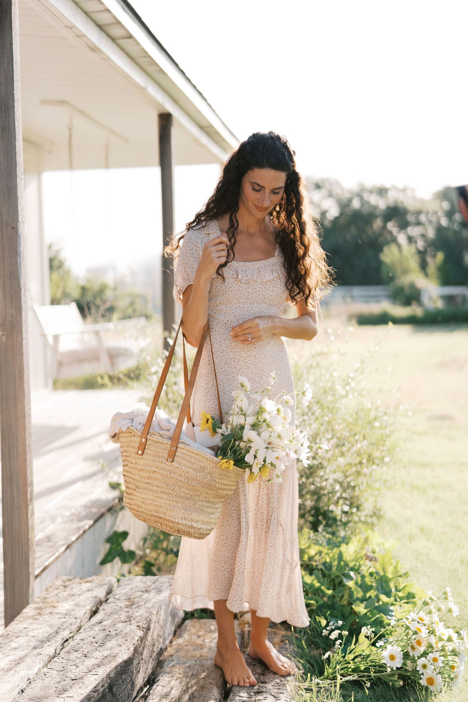 A woman in a floral dress holds a straw tote with wildflowers while standing on a porch in sunlight.