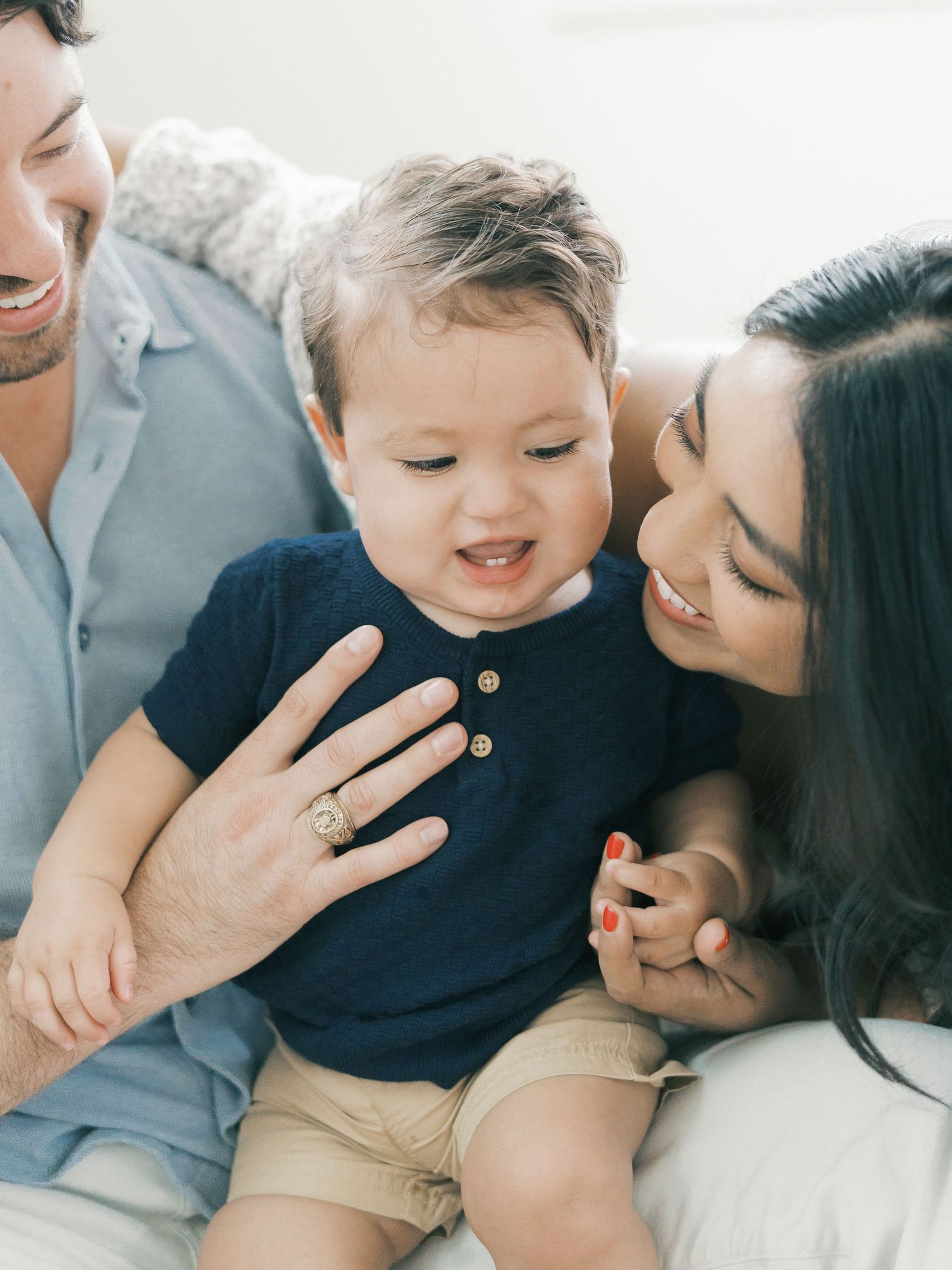 Two parents smile lovingly at their cheerful toddler boy in a navy henley shirt sitting between them.