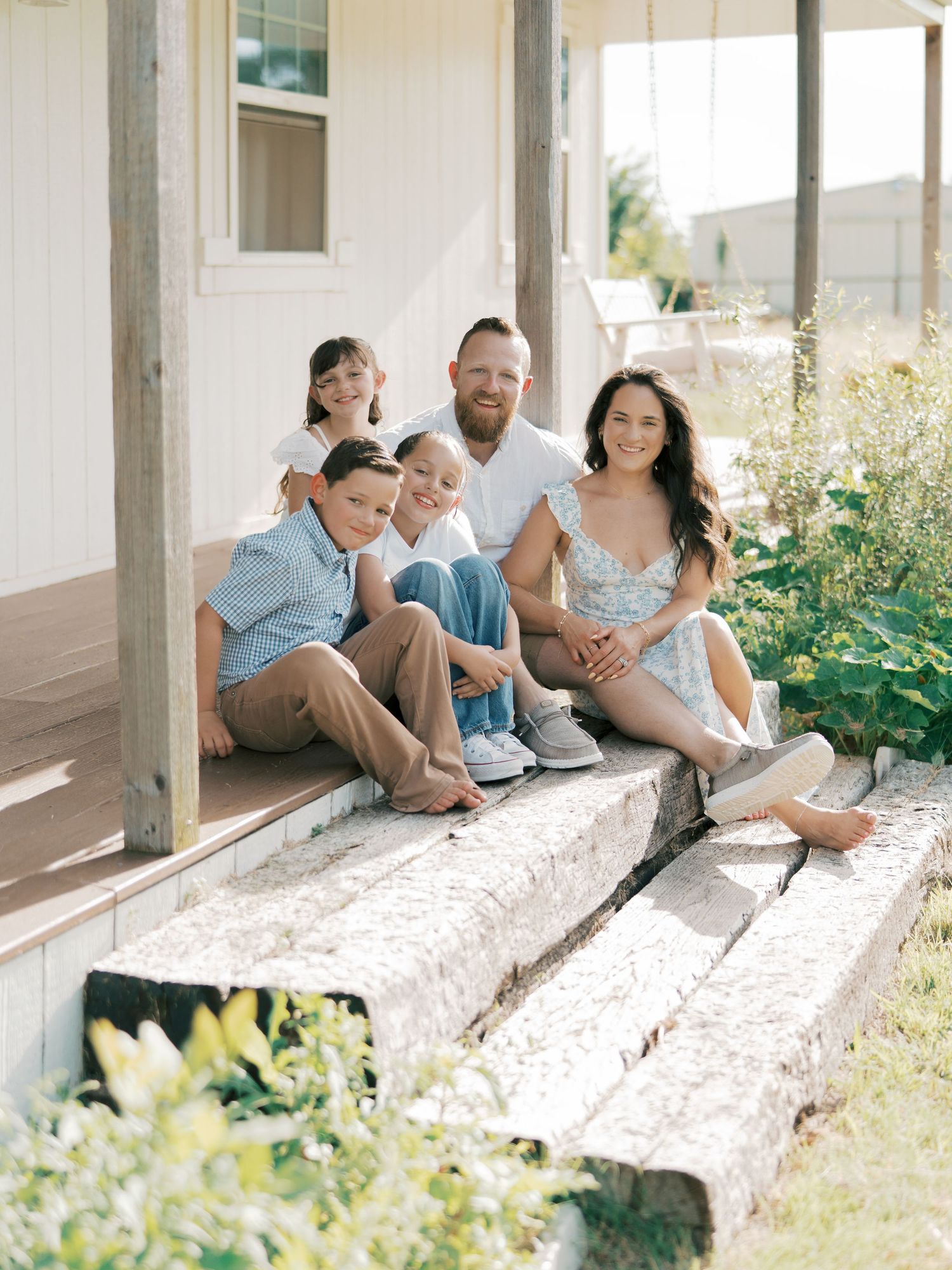 Happy family of five sitting on porch steps outside a white farmhouse by Alahir Villasenor Photographer