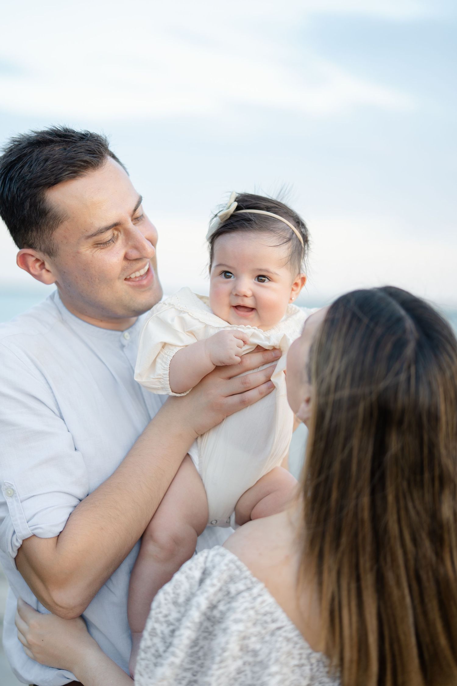 Smiling baby girl in cream outfit being held by parents at the beach on a cloudy day.