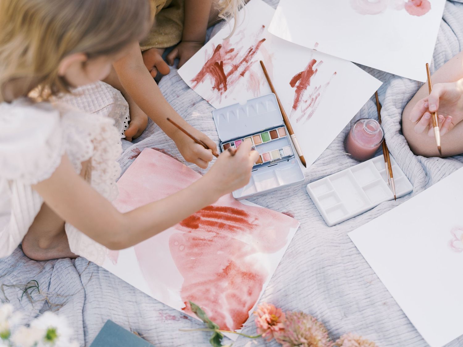 Children paint watercolor artwork on paper spread across a blanket, surrounded by flowers and art supplies.