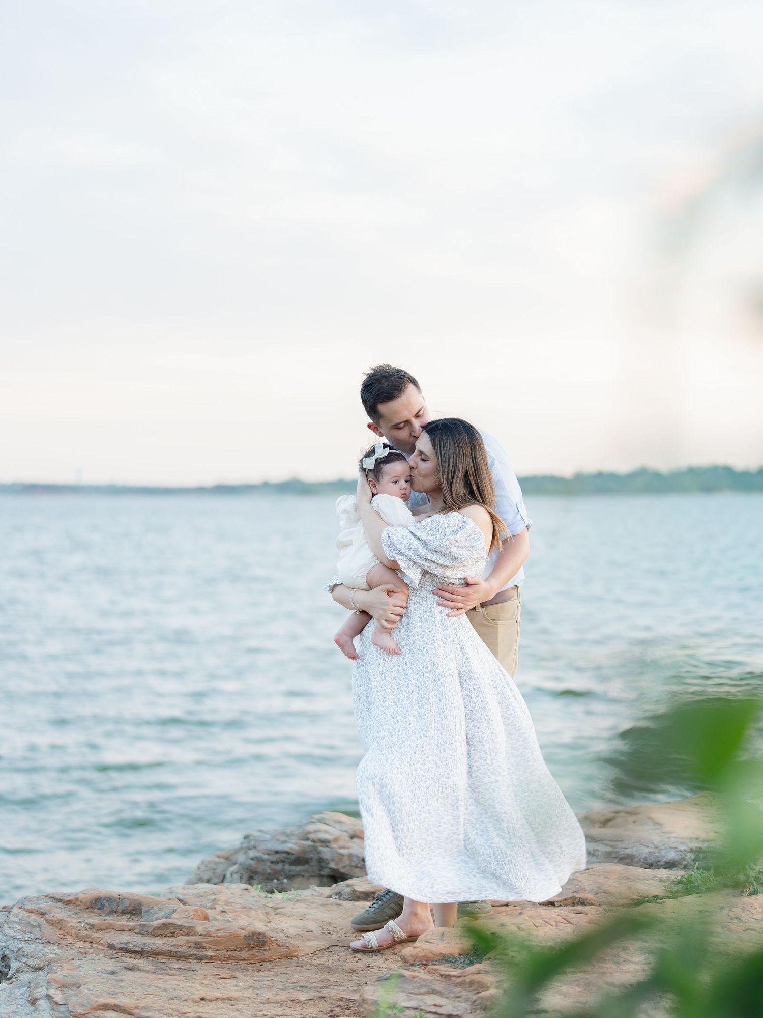 A family of three shares a tender embrace on rocky shoreline with calm turquoise water and soft sky behind them.