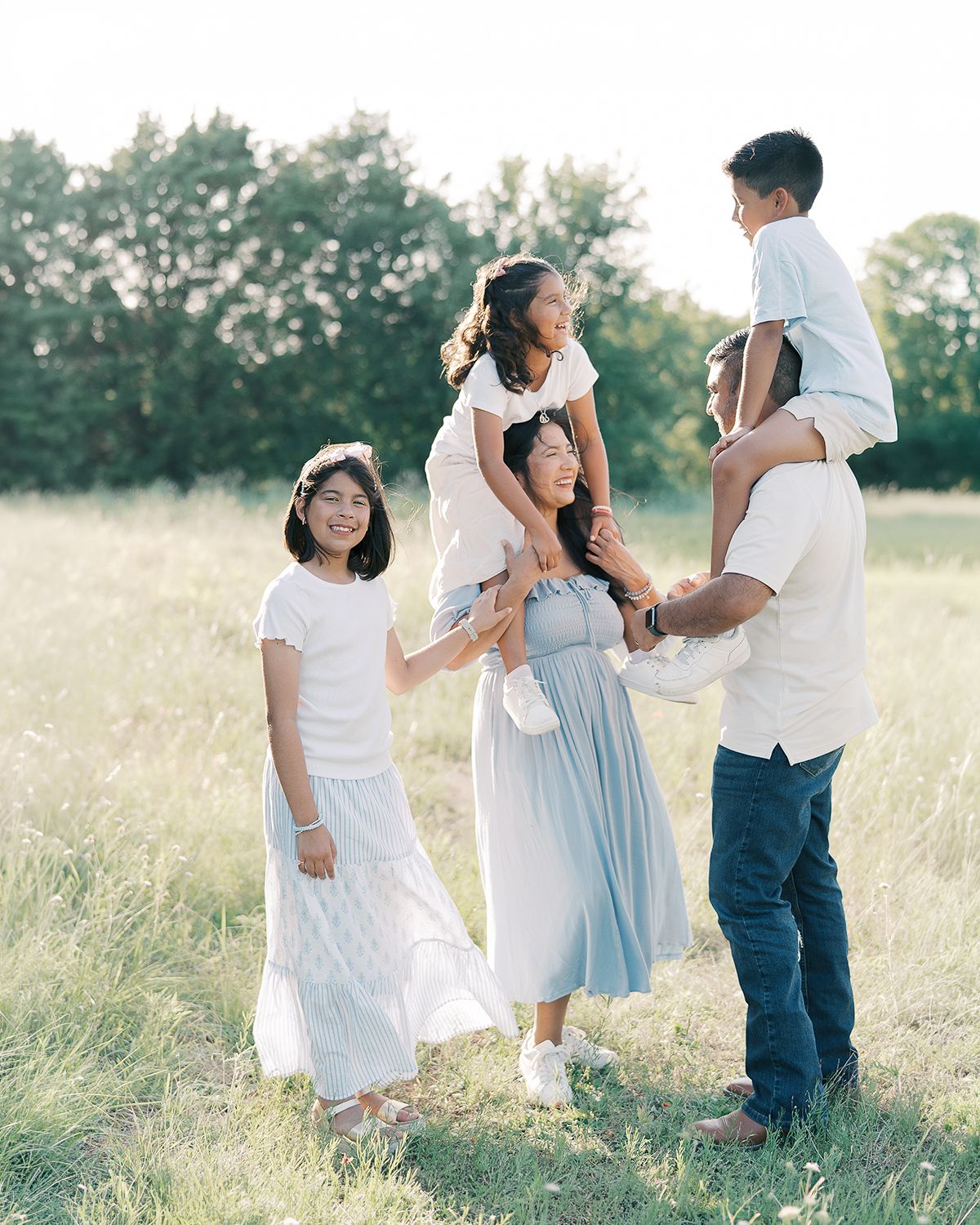 A joyful Asian family of five poses together in a sunny green meadow, with children on shoulders by Alahir Villasenor Photographer