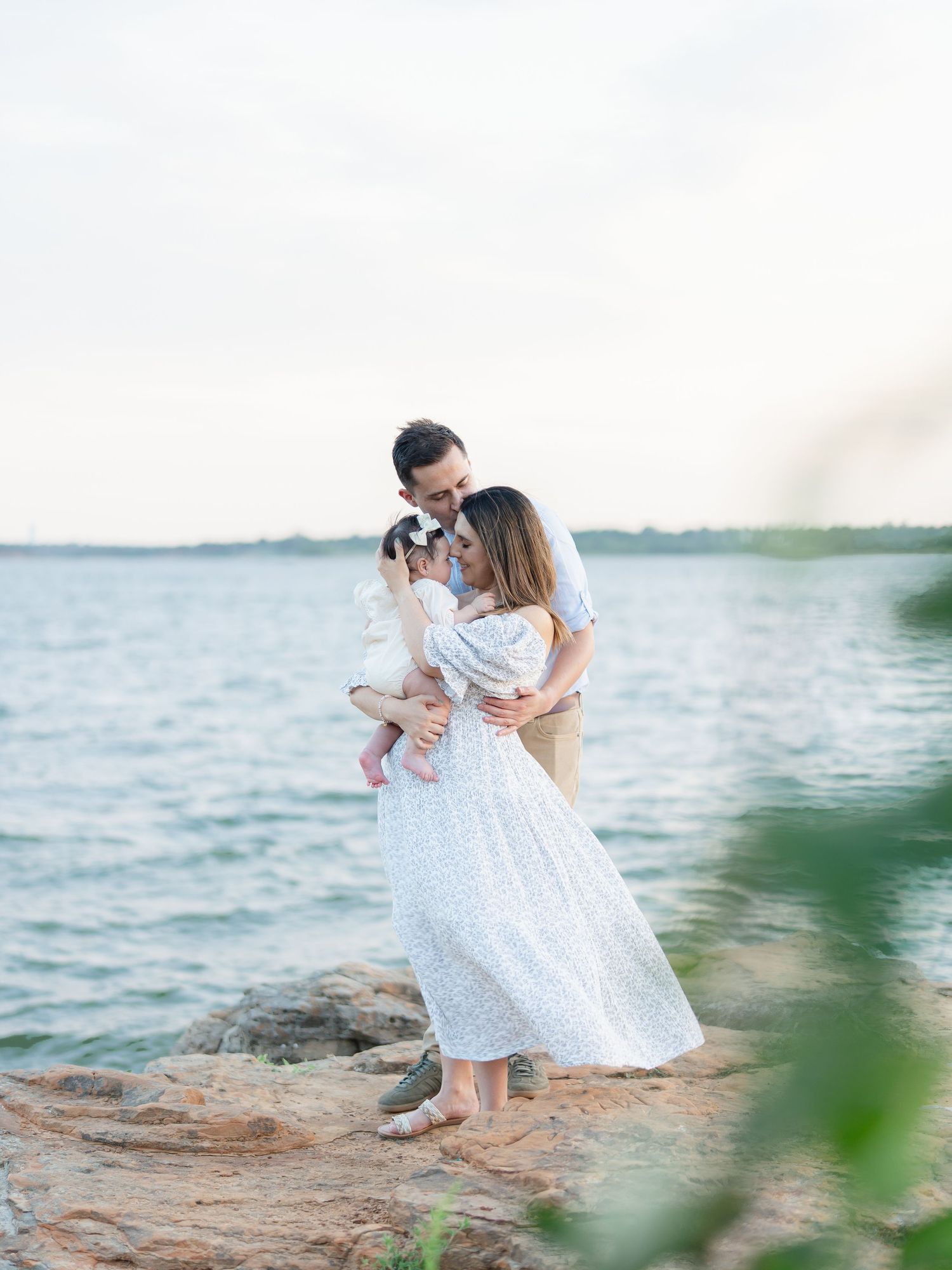 Parents in white outfits cuddling baby girl on rocky shore with ocean backdrop at dusk.