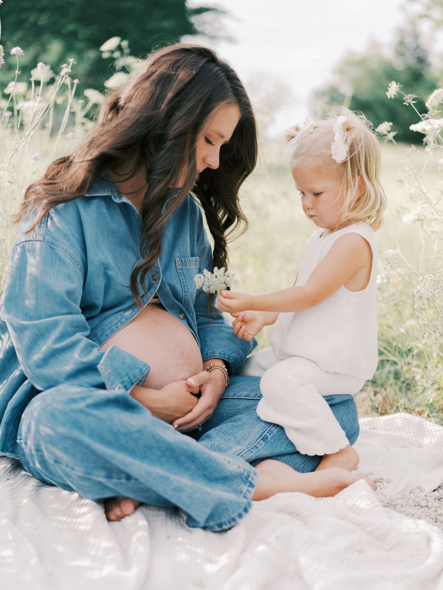 A pregnant woman in denim sits outdoors with her toddler daughter who offers her a wildflower in a sunny field.